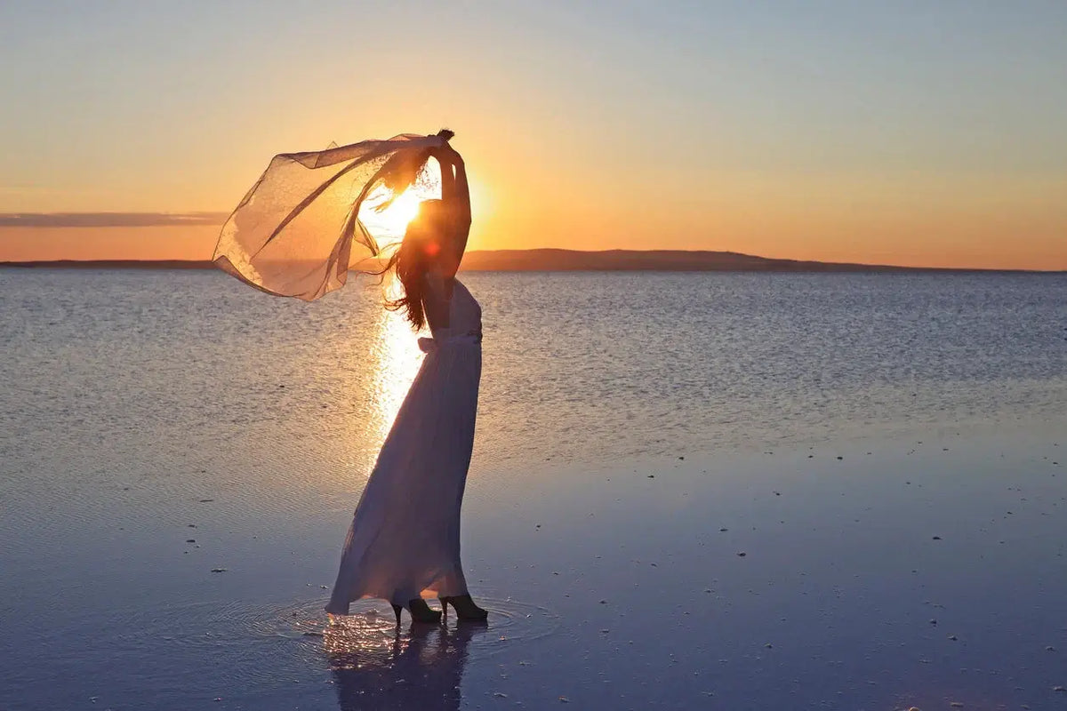 Femme dans une robe blanche élégante tenant un voile devant un coucher de soleil, représentant l'équipements mode femme élégante.