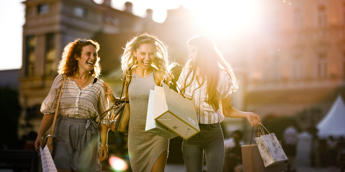 Trois femmes souriantes faisant du shopping au soleil, profitant des offres de mode femme outlet.