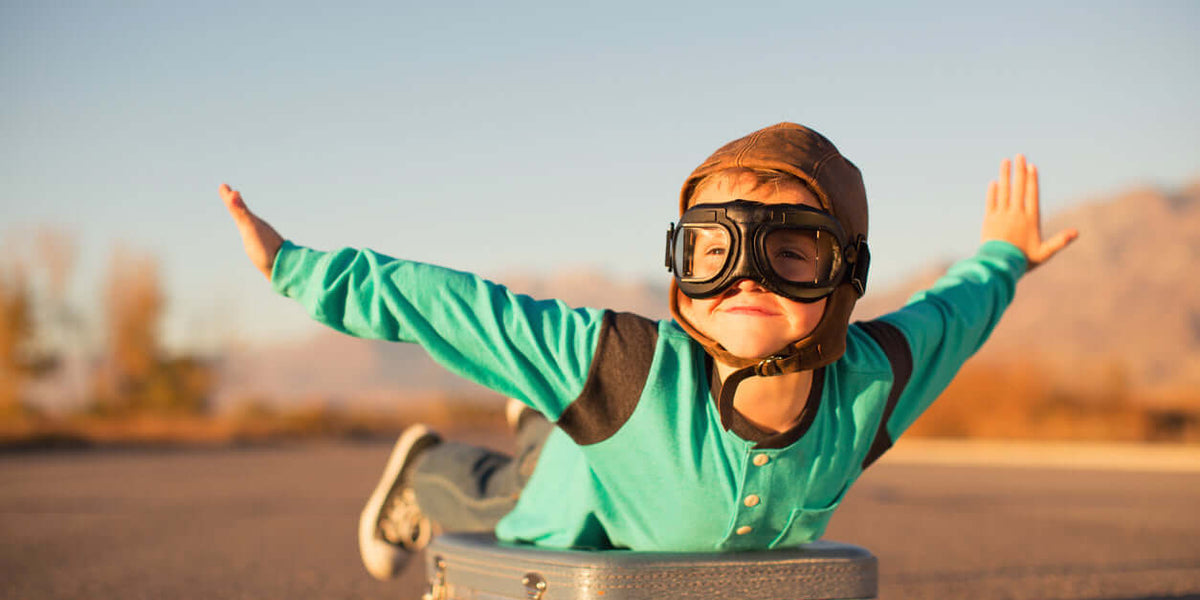 A child pretending to fly on a suitcase, wearing stylish children's accessories like goggles and a cap.