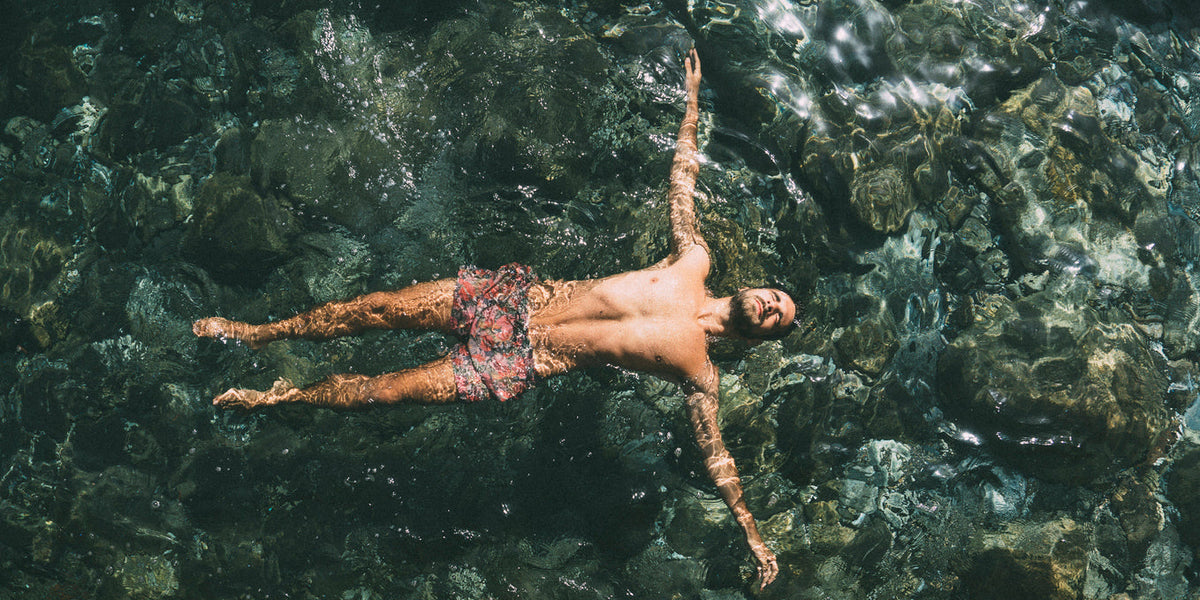 Homme flottant dans l'eau, portant des maillots de bain homme élégants, entouré de rochers et d'eau claire.