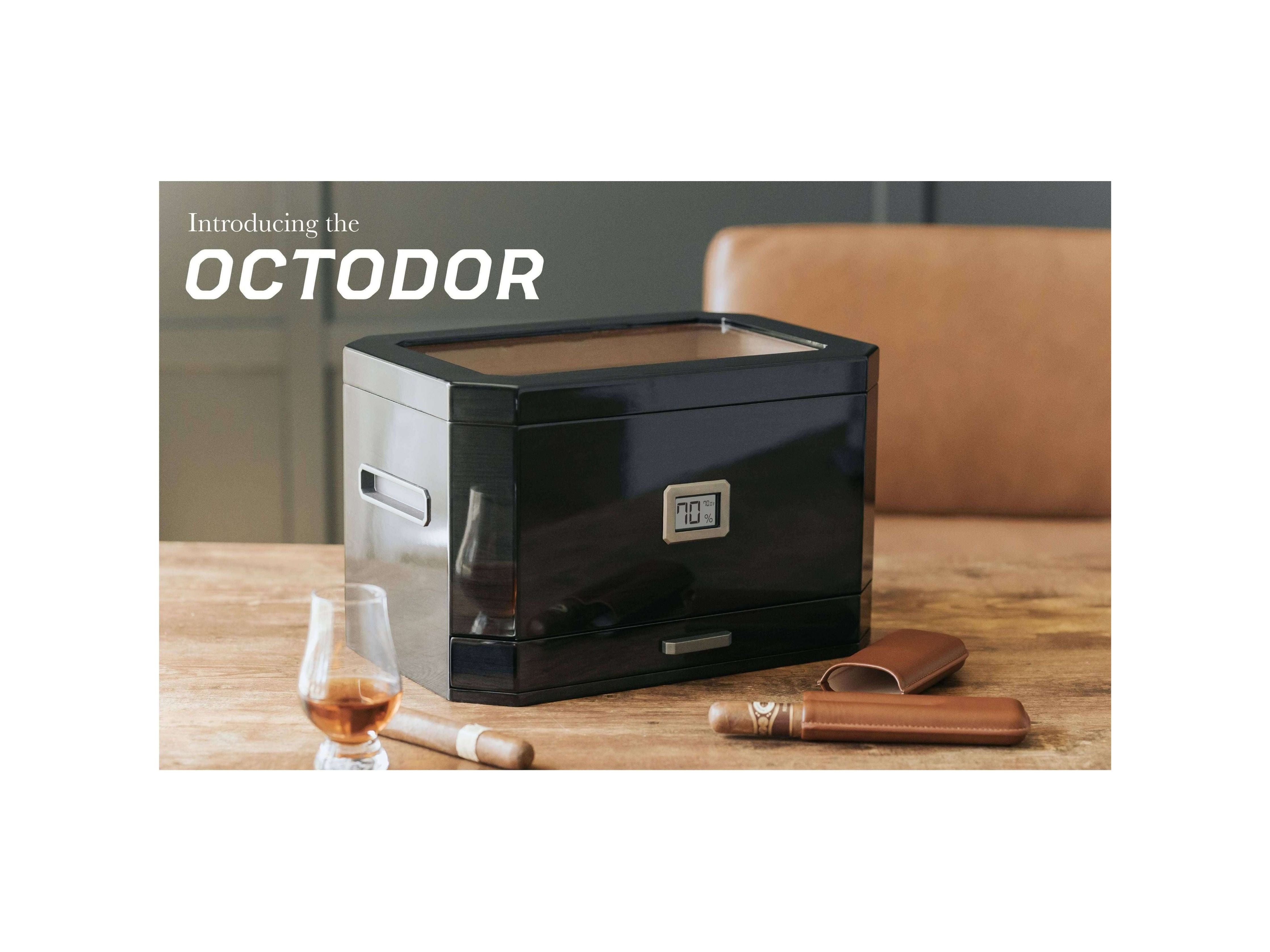 Humidor à couvercle en verre Octodor de Klaro sur une table, avec cigares et un verre de whisky.