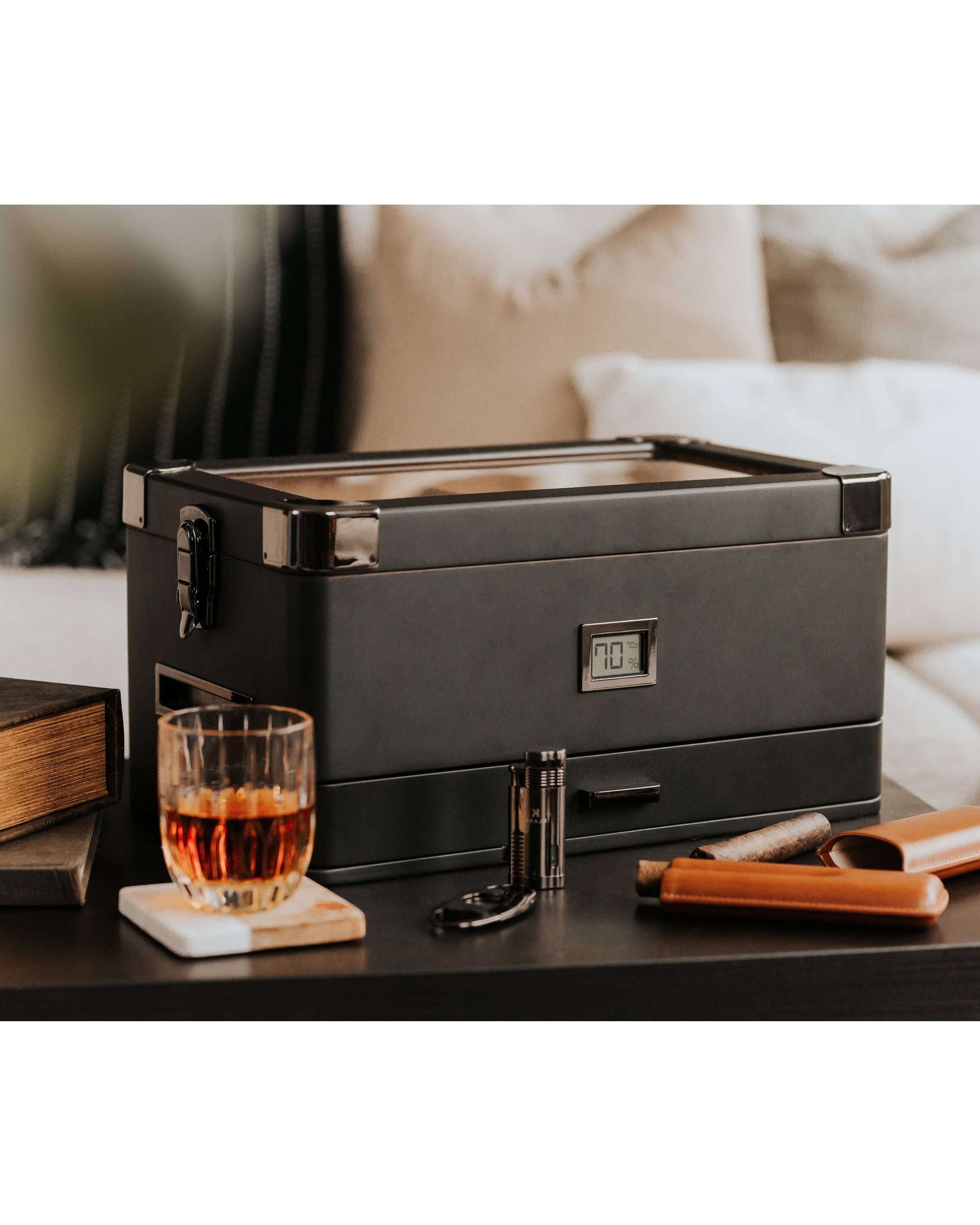 Humidor militaire Black Edition with black matte finish, next to a glass of whiskey and cigar accessories on a table.