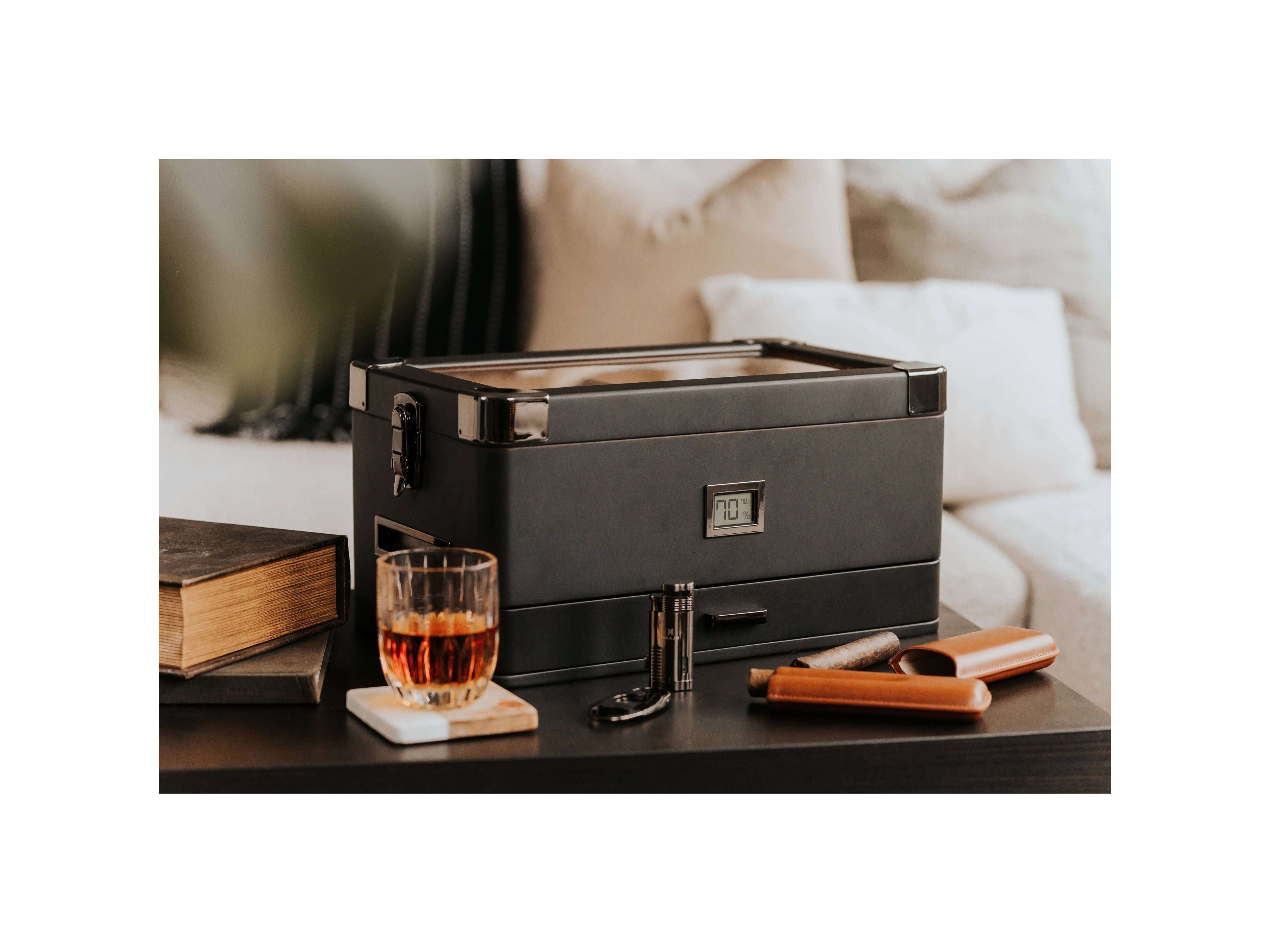 Humidor militaire Black Edition with black matte finish, next to a glass of whiskey and cigar accessories on a table.