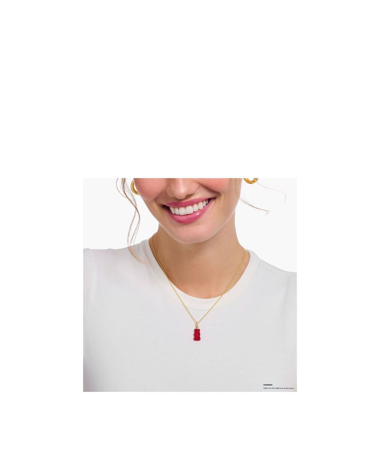 Femme souriante portant une chaîne dorée avec une breloque ours gélifié rouge en cristal, sur un t-shirt blanc.