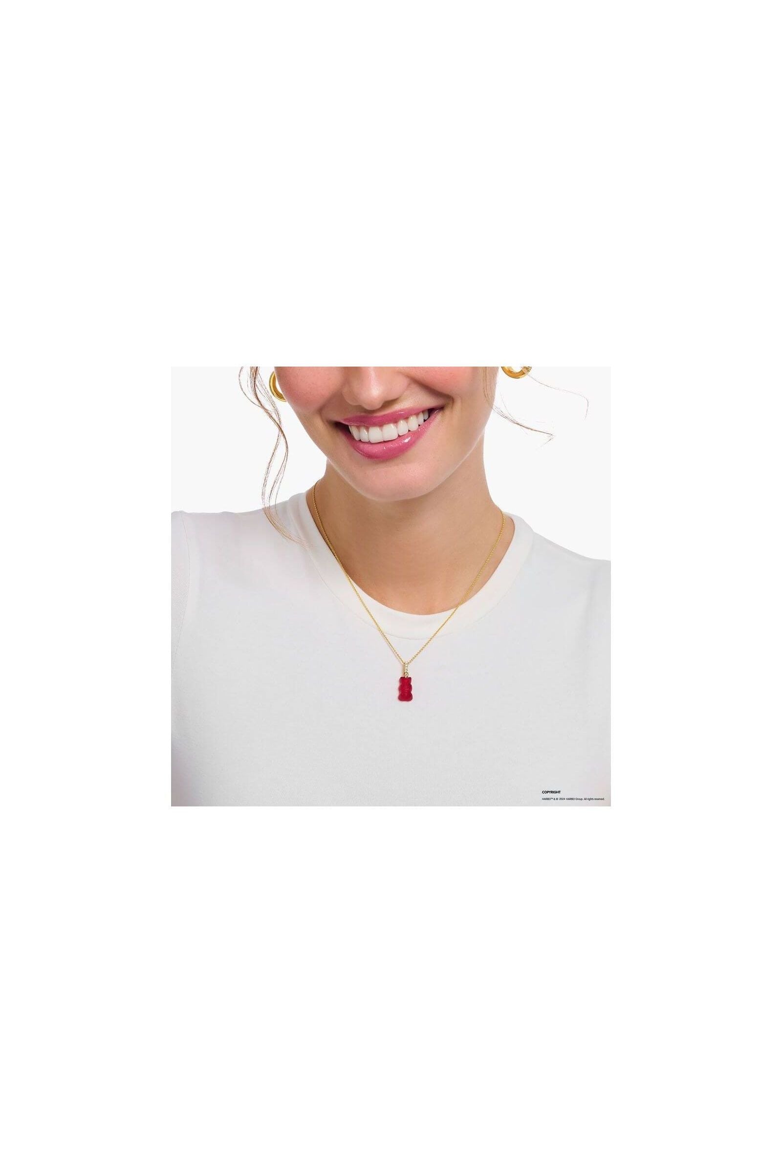 Femme souriante portant une chaîne dorée avec une breloque ours gélifié rouge en cristal, sur un t-shirt blanc.