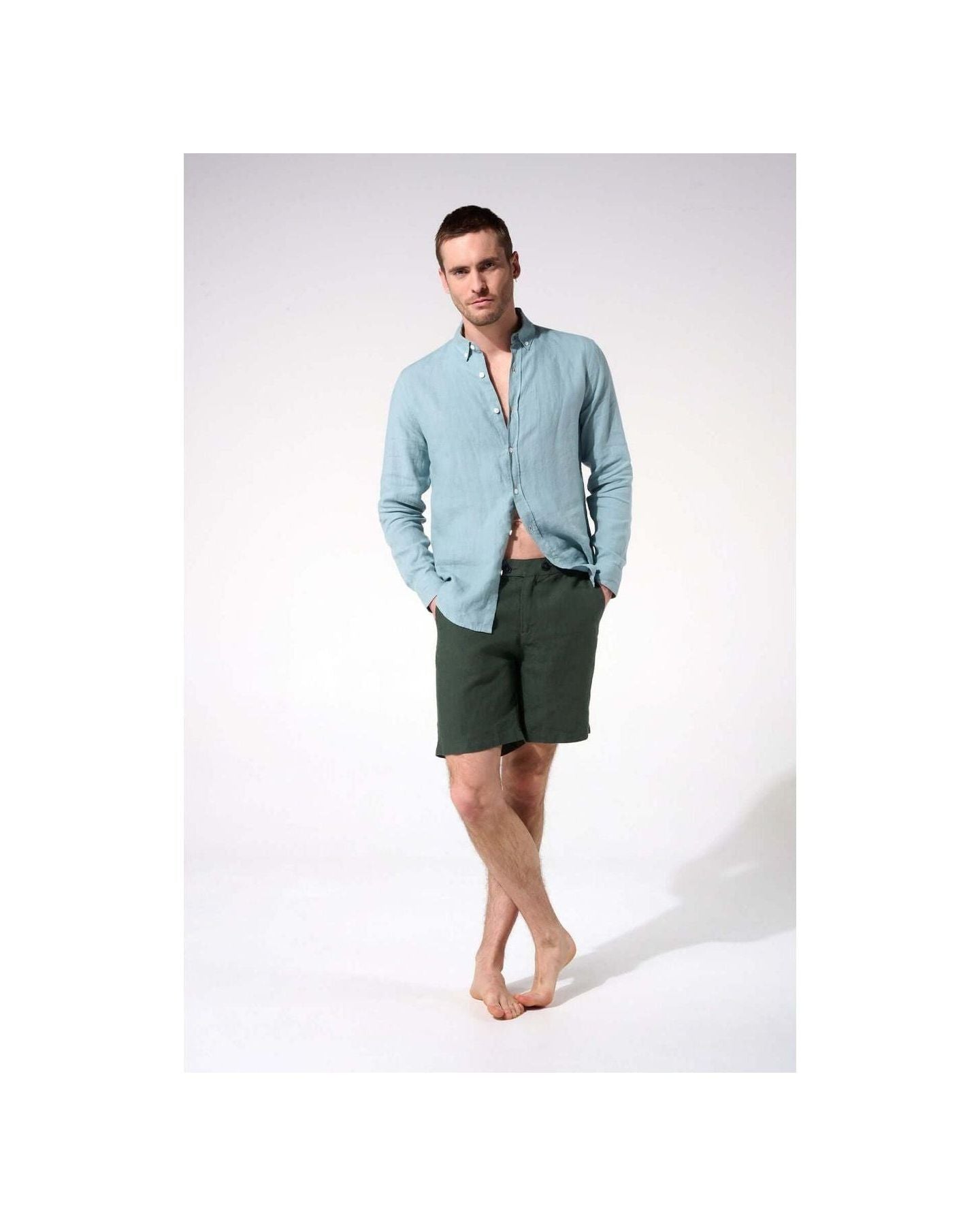 Man wearing a light blue linen shirt and dark green shorts, standing barefoot in a casual pose.