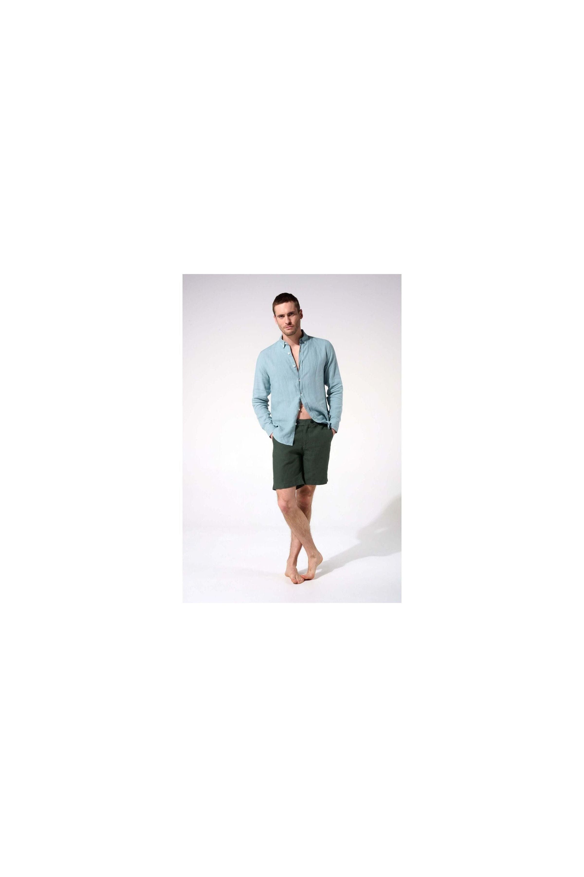 Man wearing a light blue linen shirt and dark green shorts, standing barefoot in a casual pose.