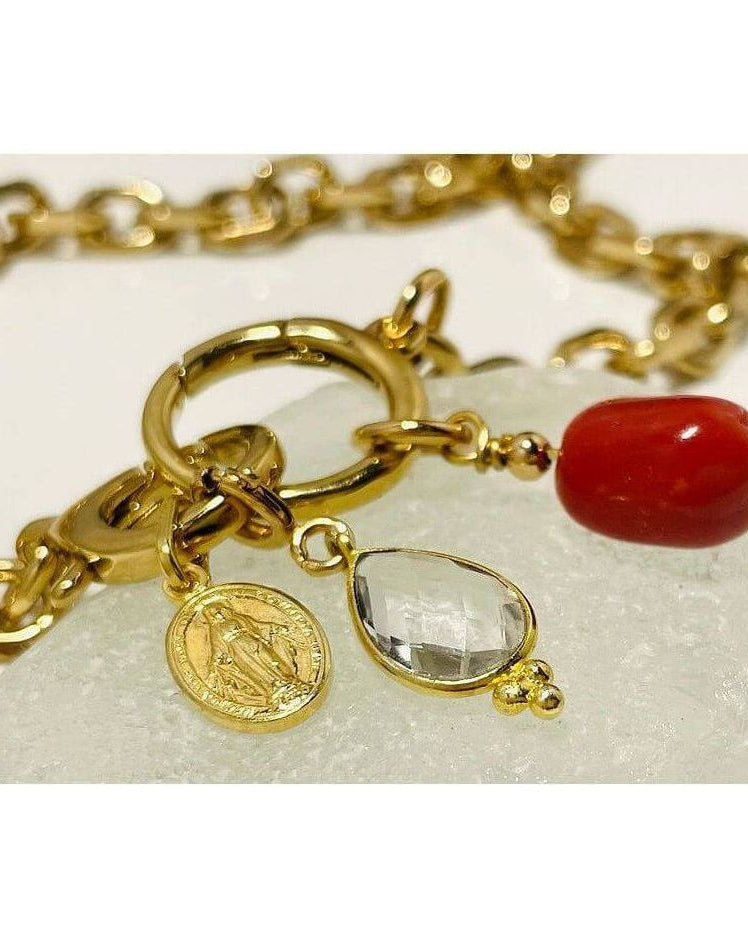 Collier Madone bracelet featuring gold links, red coral bead, and a clear gem on a light stone background.