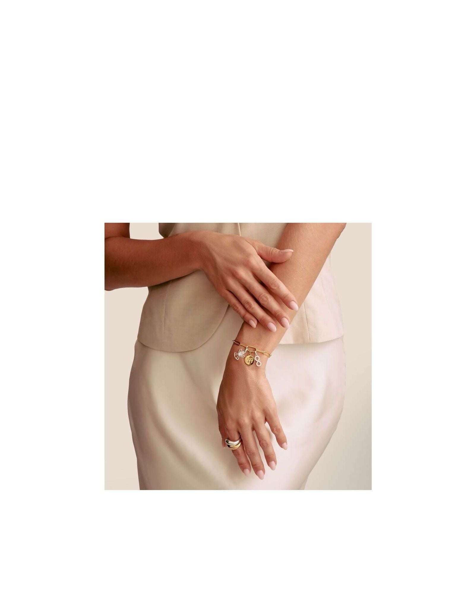 Modeled hand wearing a gold charm bracelet and rings, showcasing stylish accessories against a beige background.