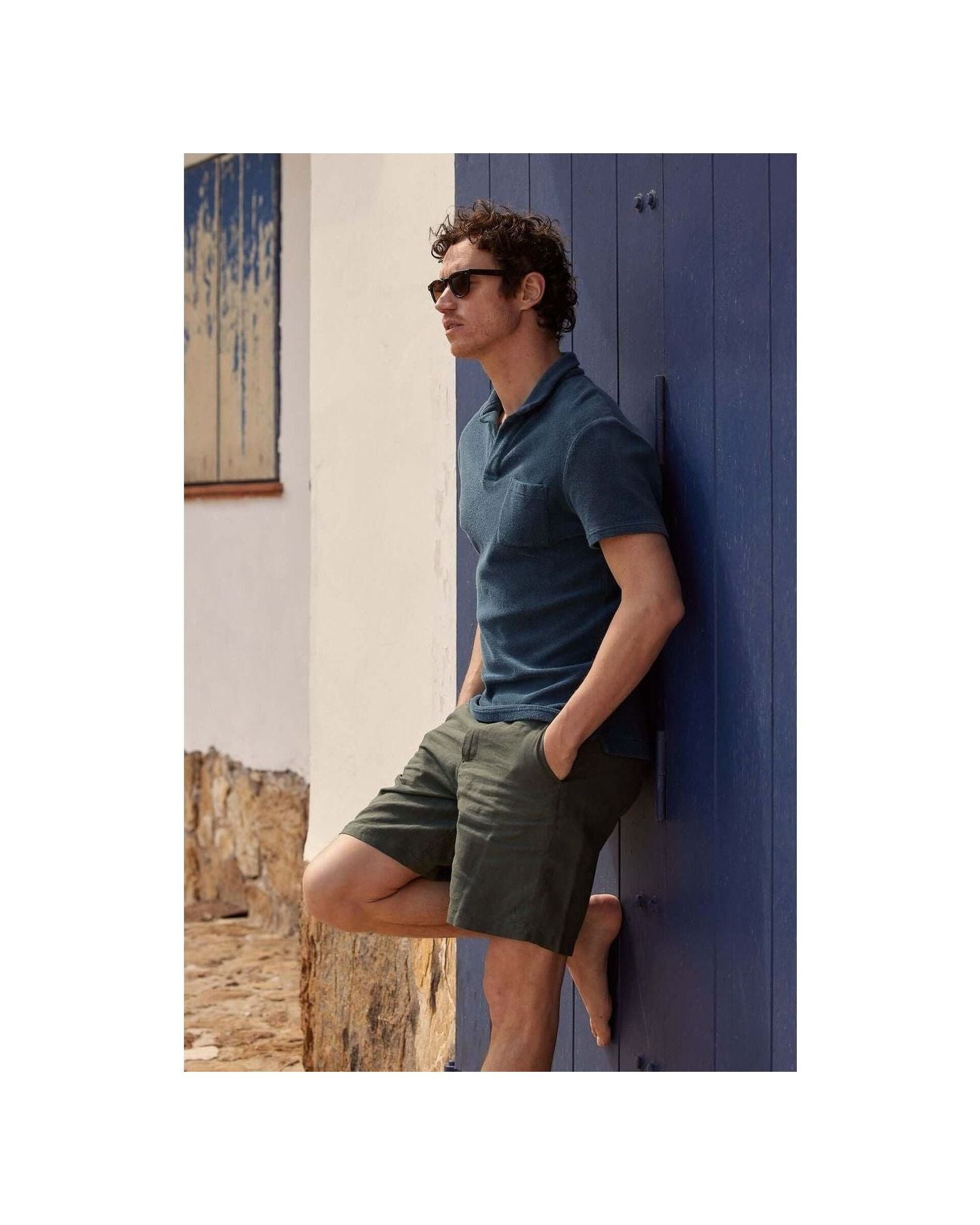 Man wearing Bermuda lin Balsam shorts and a polo shirt, leaning against a blue wall in a stylish summer look.