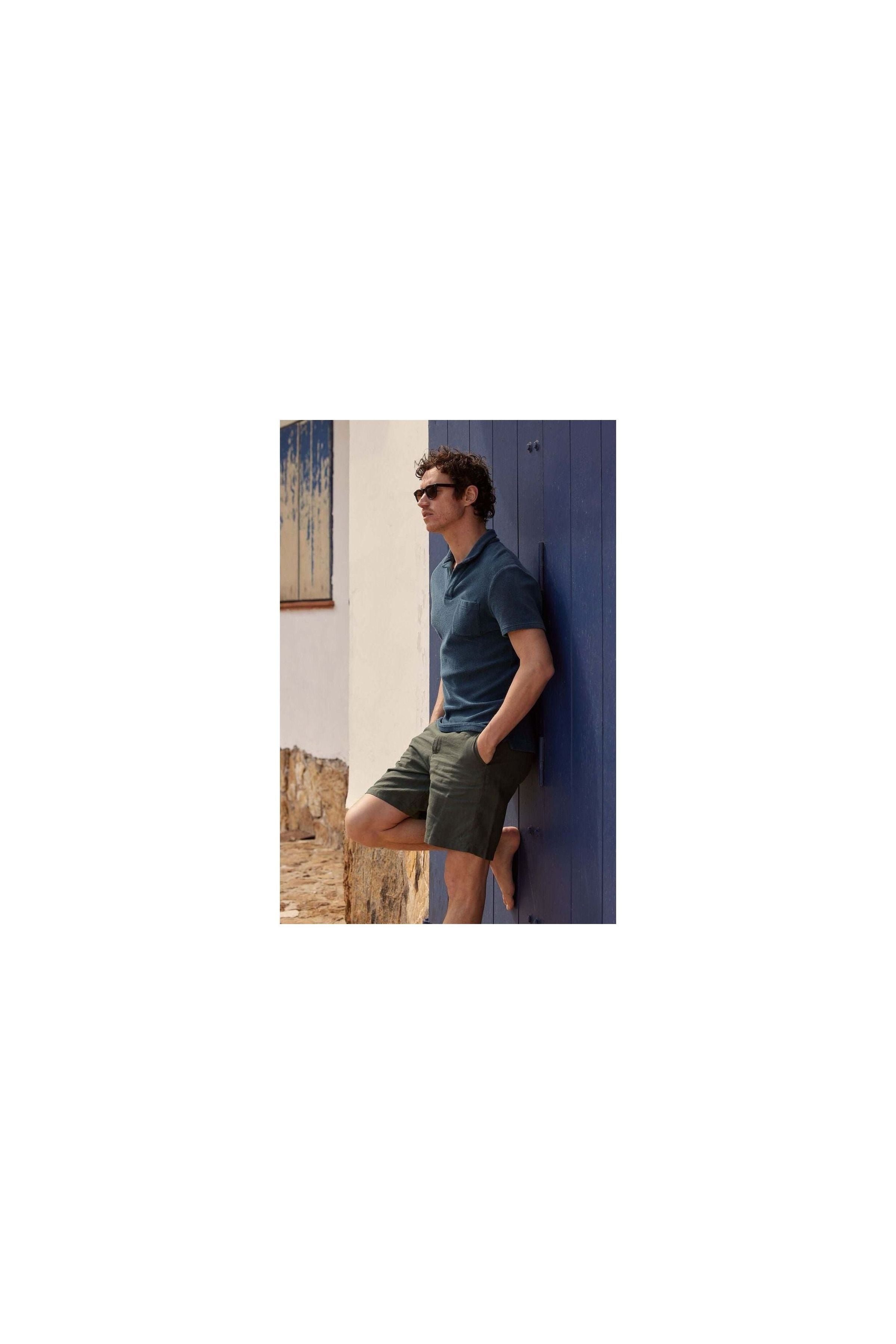 Man wearing Bermuda lin Balsam shorts and a polo shirt, leaning against a blue wall in a stylish summer look.