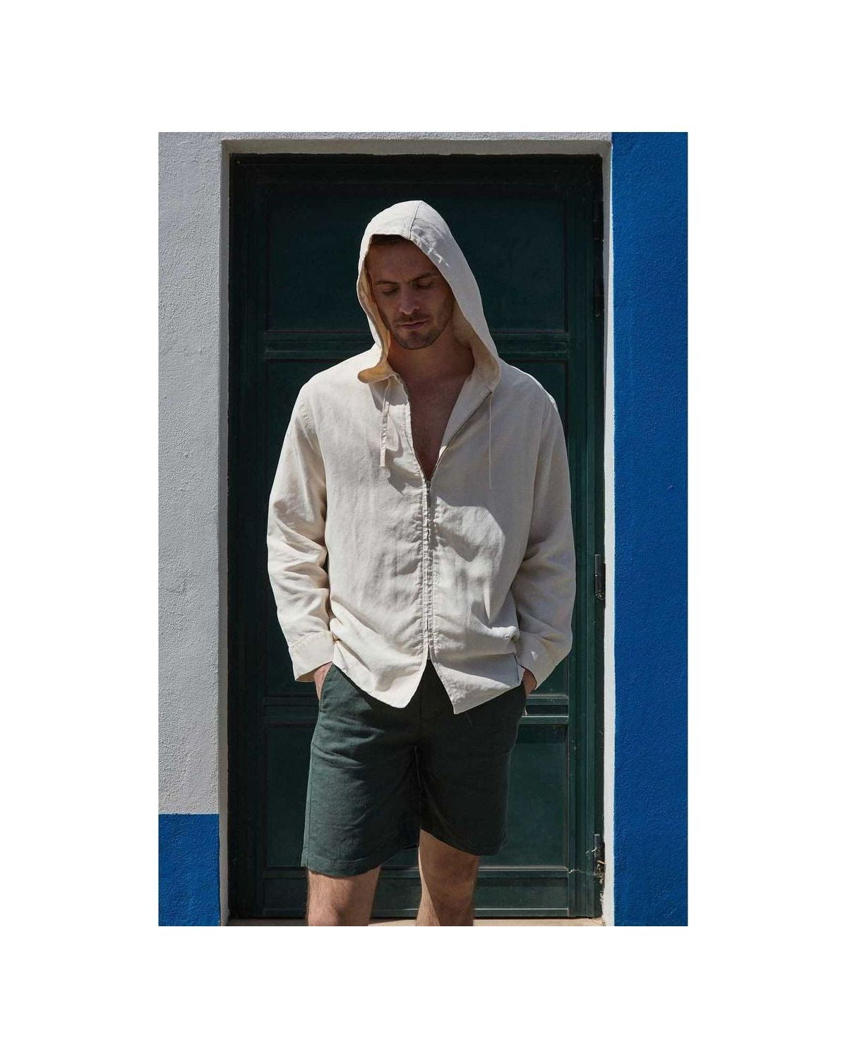A man wearing a light beige hooded shirt and dark shorts stands in front of a blue and white wall.