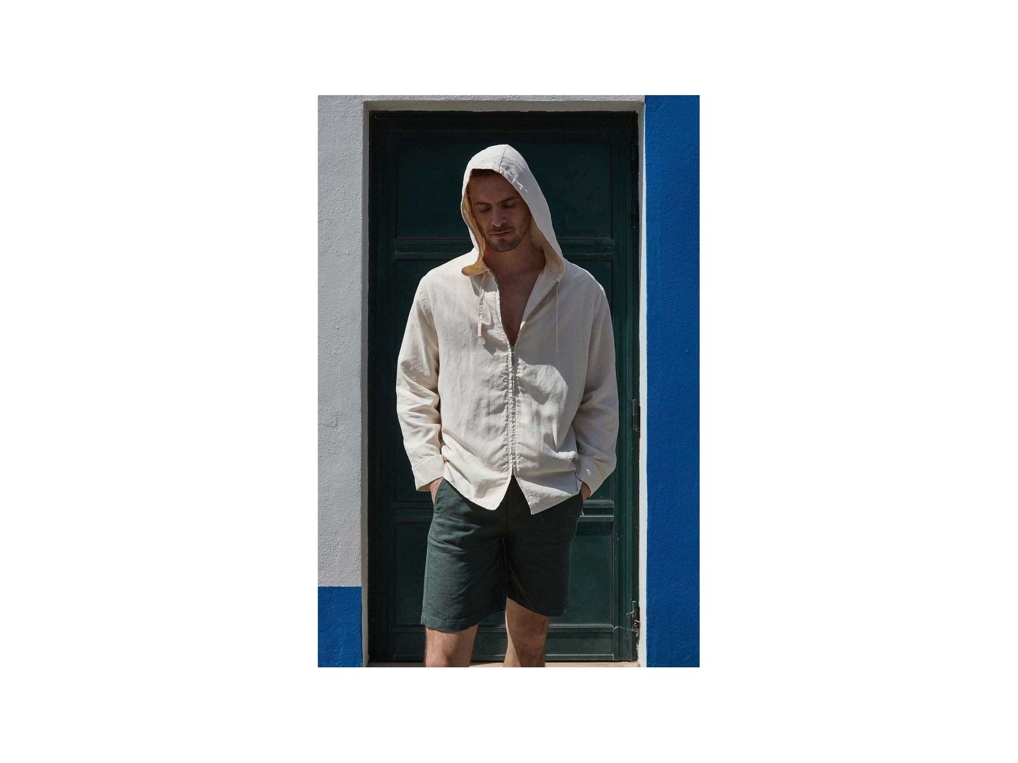 A man wearing a light beige hooded shirt and dark shorts stands in front of a blue and white wall.
