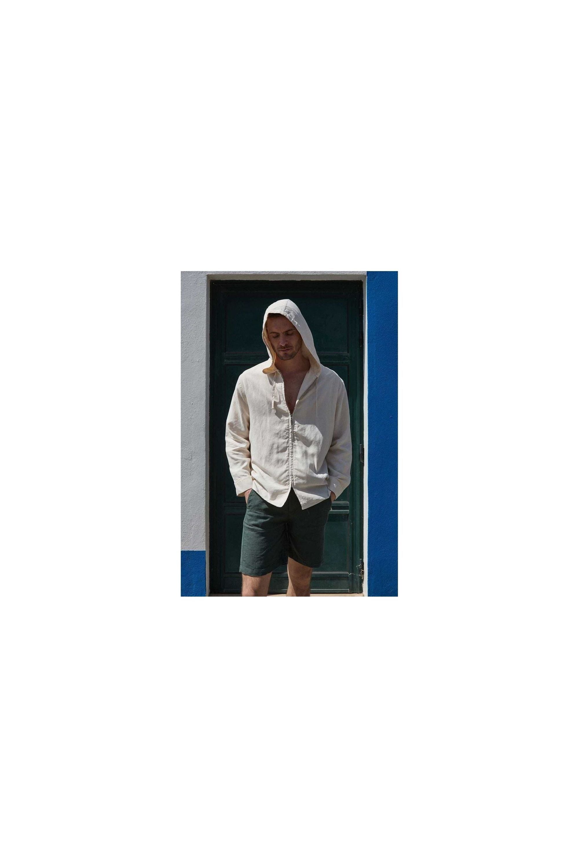 A man wearing a light beige hooded shirt and dark shorts stands in front of a blue and white wall.
