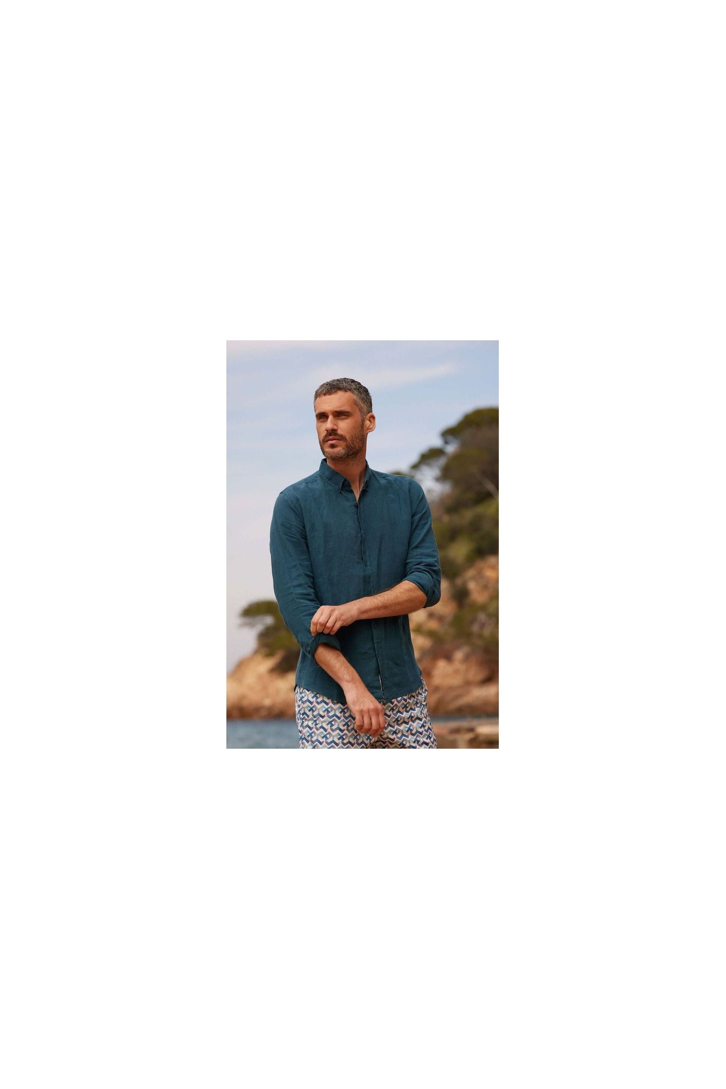 Homme avec chemise en lin bleu, col boutonné, décor naturel, style décontracté en bord de mer.