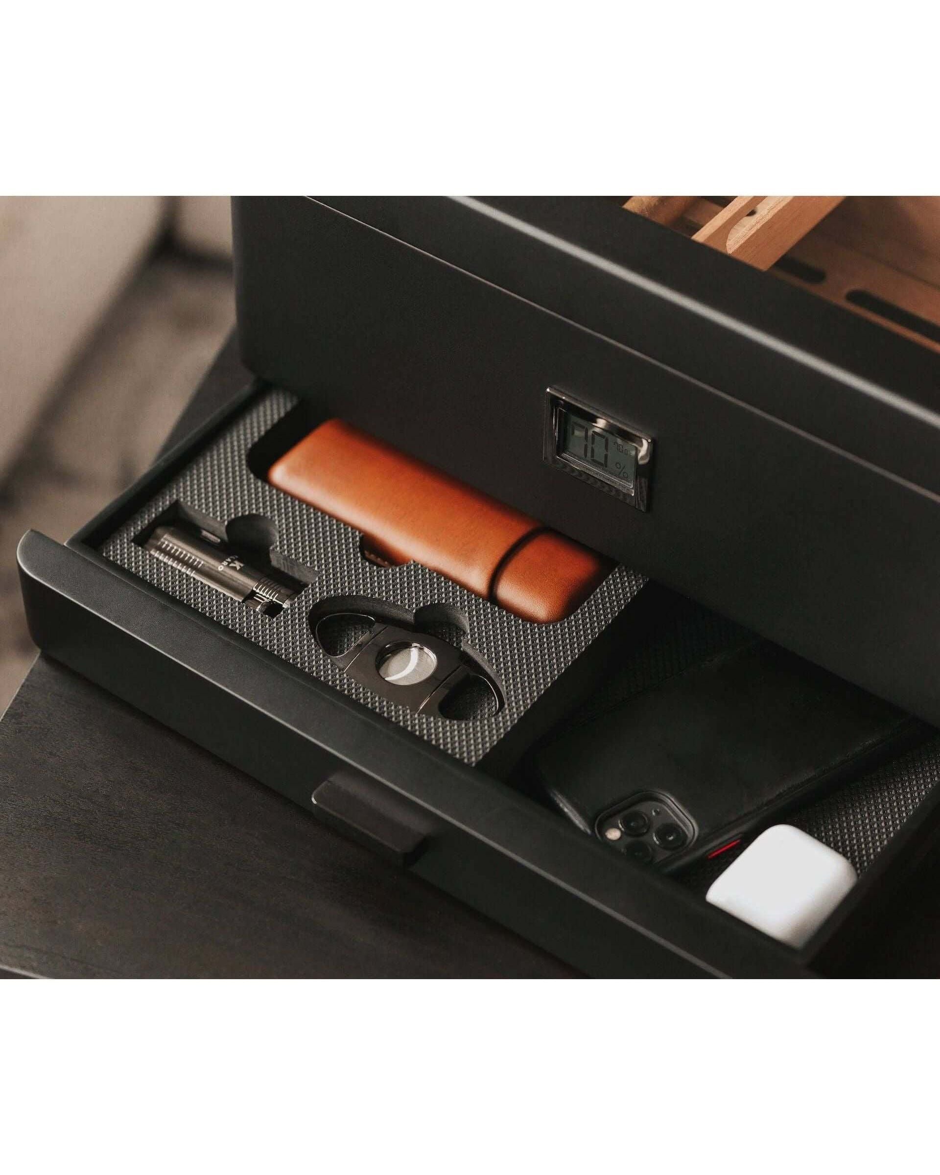 Humidor militaire Black Edition open drawer showcasing accessories like a cigar cutter, leather case, and smartphone.