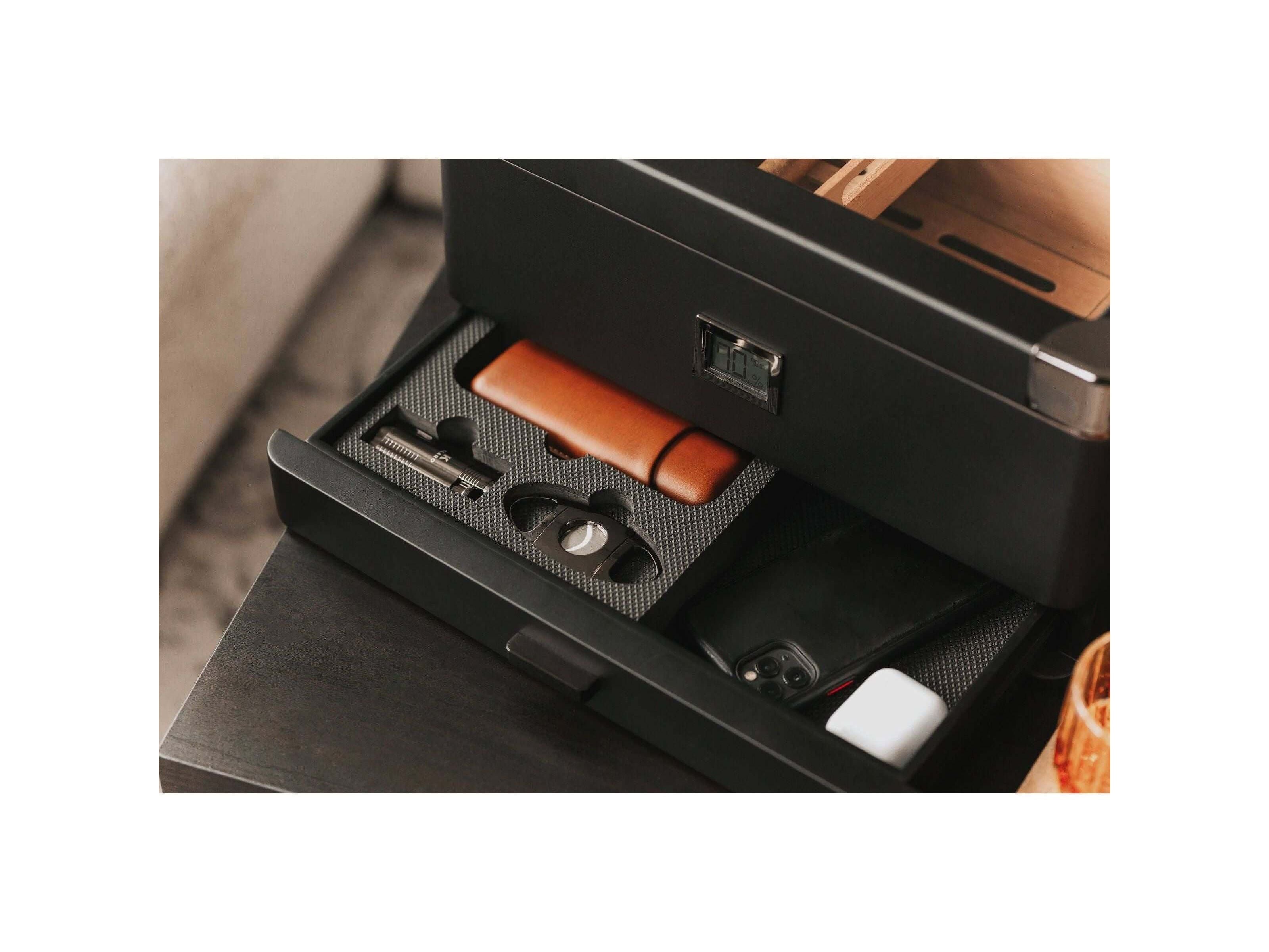 Humidor militaire Black Edition open drawer showcasing accessories like a cigar cutter, leather case, and smartphone.