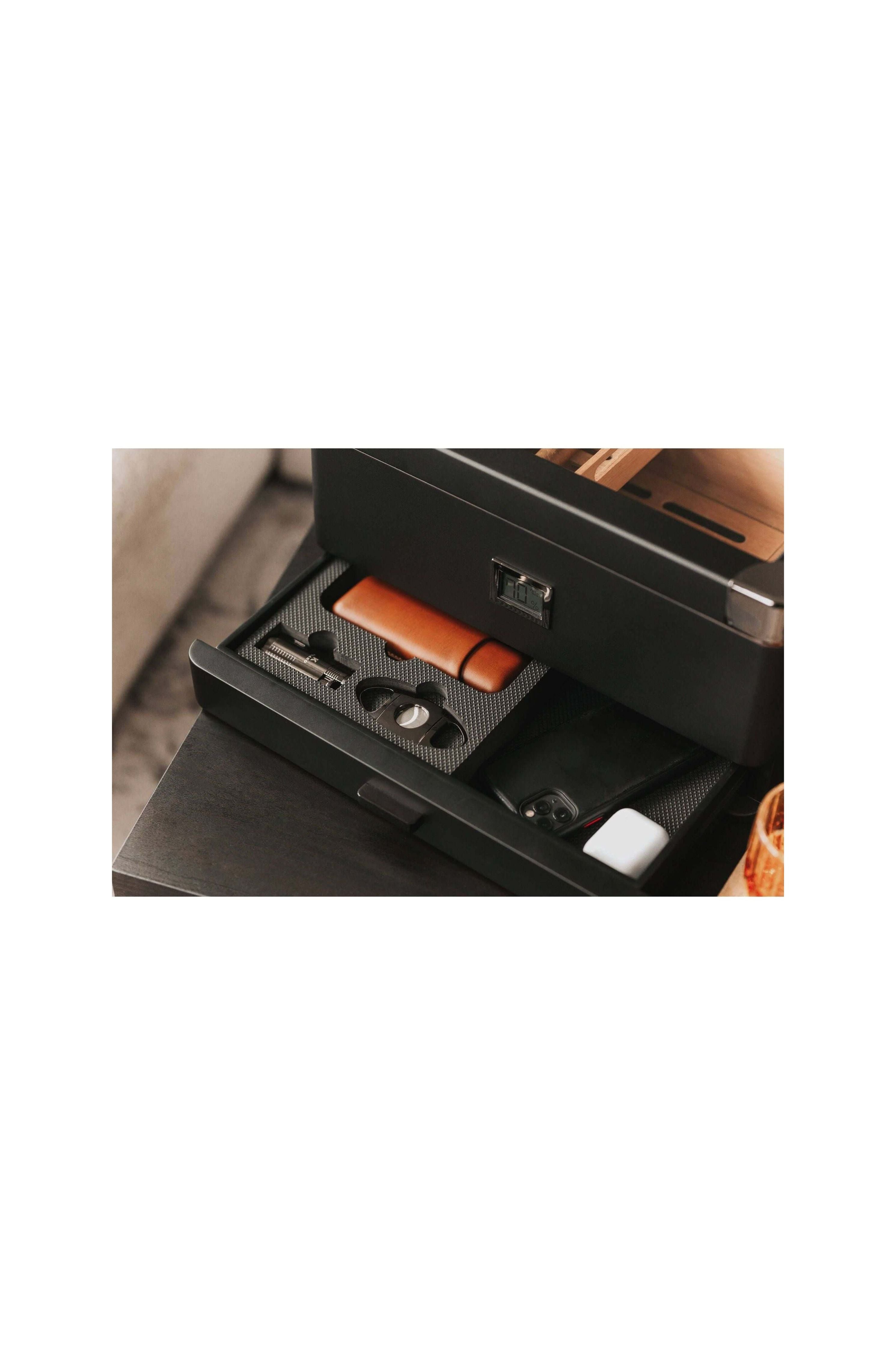 Humidor militaire Black Edition open drawer showcasing accessories like a cigar cutter, leather case, and smartphone.