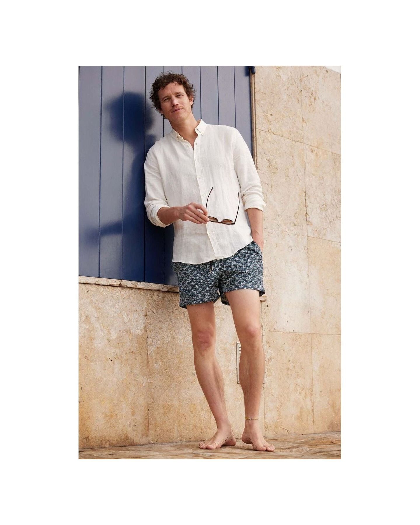 Homme en short de bain Ecume, style décontracté avec chemise blanche, posant devant un mur beige.