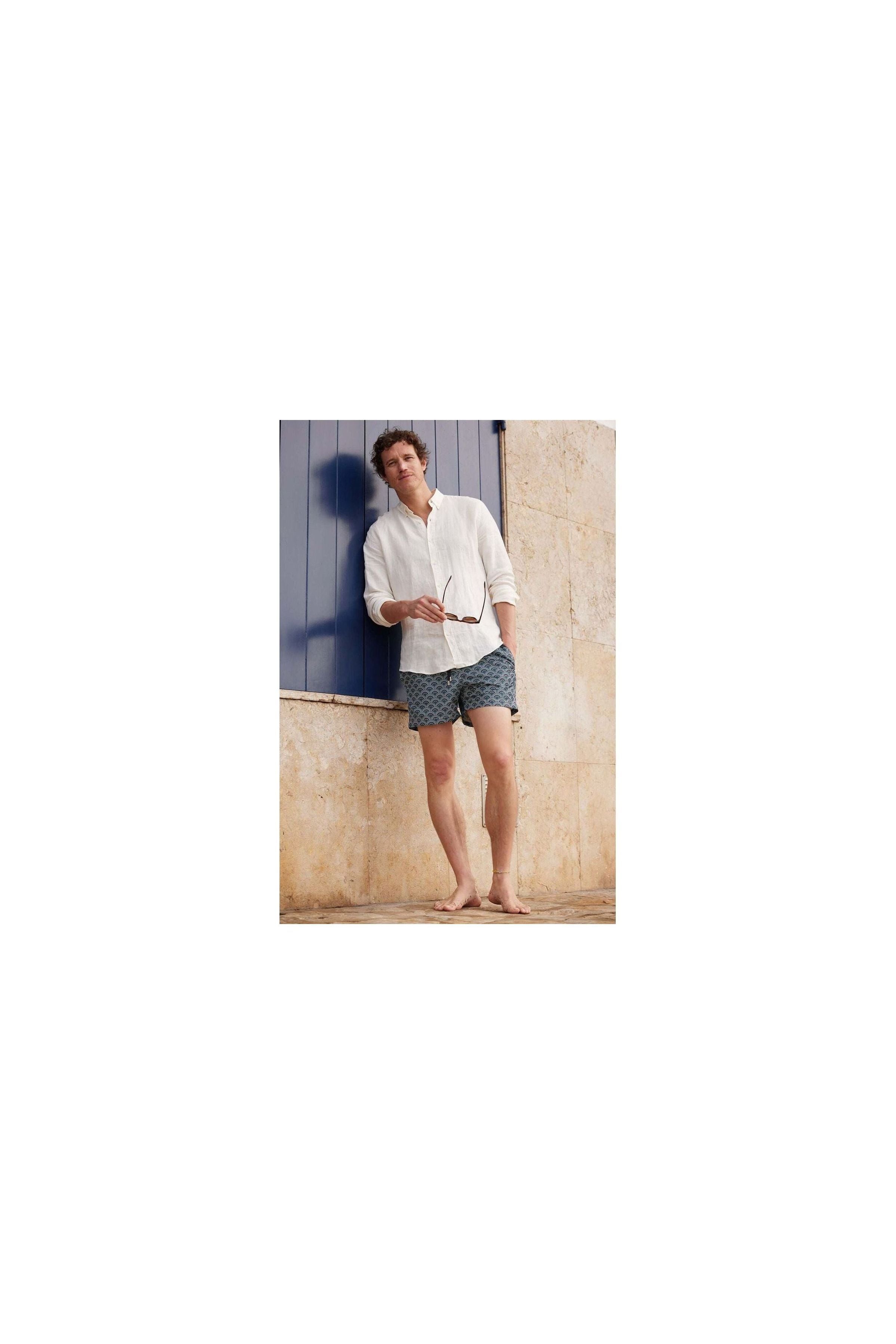 Homme en short de bain Ecume, style décontracté avec chemise blanche, posant devant un mur beige.