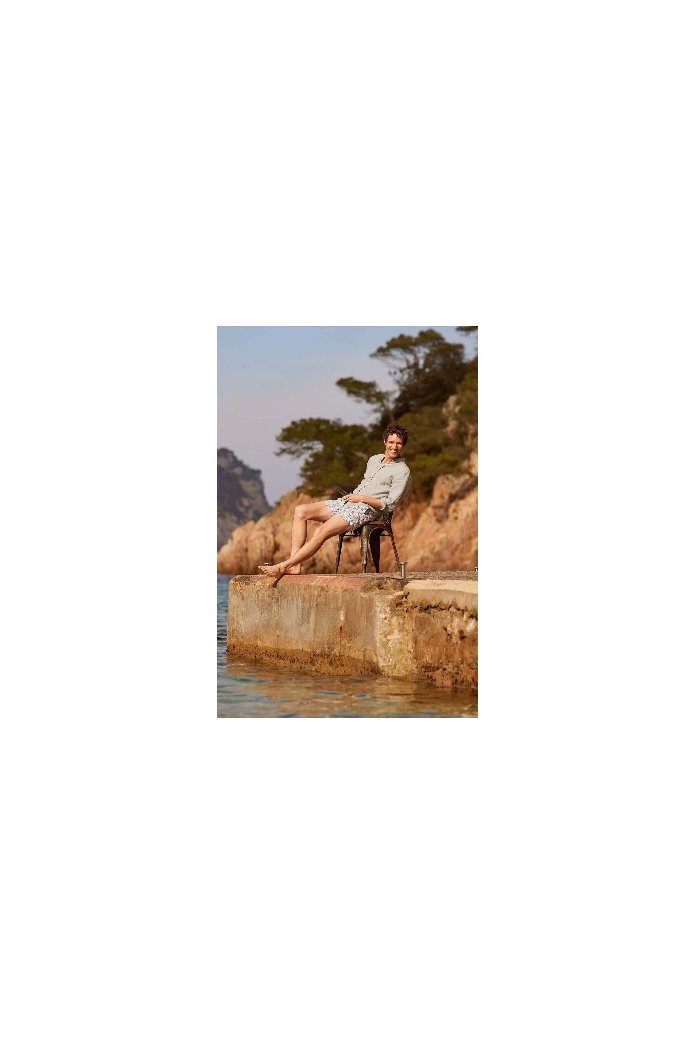 Homme détendu en short de bain Cannage assis sur un quai, en bord de mer, avec des montagnes en arrière-plan.