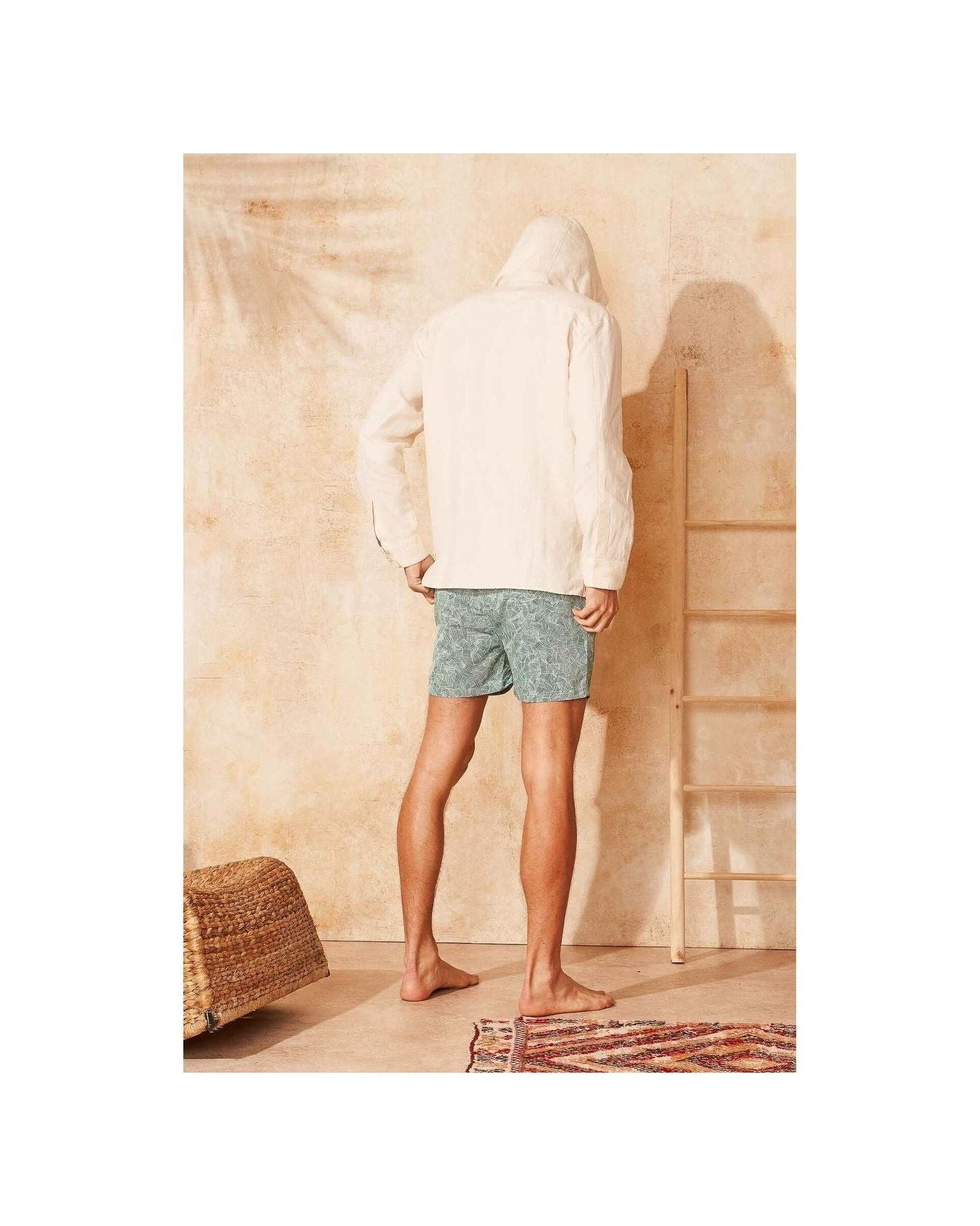 Homme vu de dos portant des shorts de bain Reflets en tissu recyclé, devant un mur beige, avec des pieds nus.