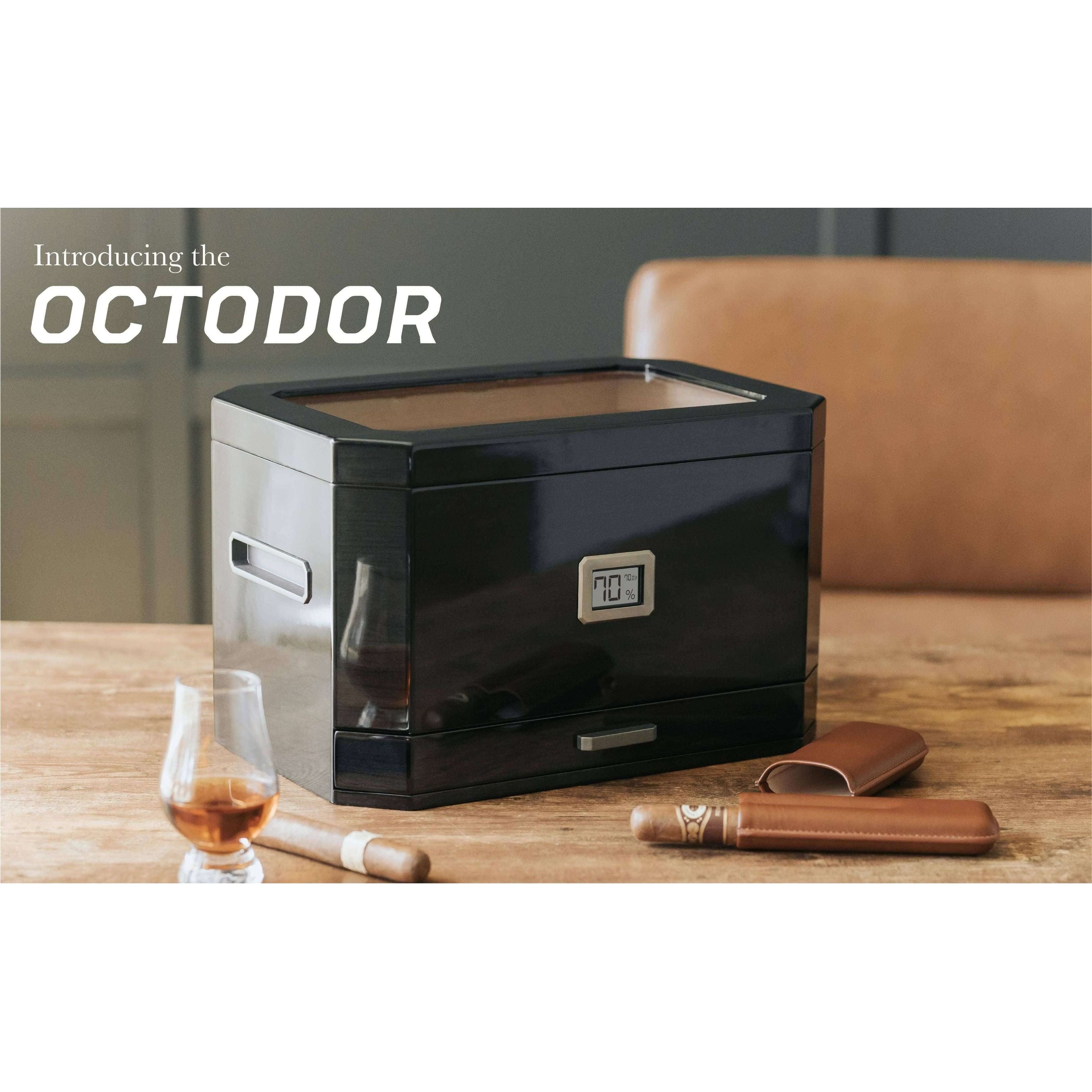 Humidor à couvercle en verre Octodor de Klaro sur une table, avec cigares et un verre de whisky.