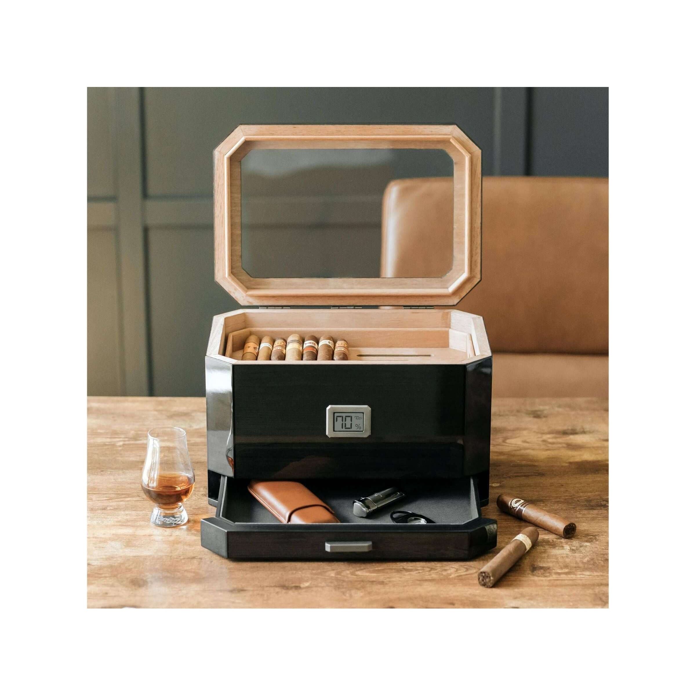 Humidor à couvercle en verre Octodor avec cigares, verre à whisky et accessoires sur une table en bois.