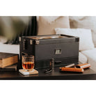 Humidor militaire Black Edition with black matte finish, next to a glass of whiskey and cigar accessories on a table.