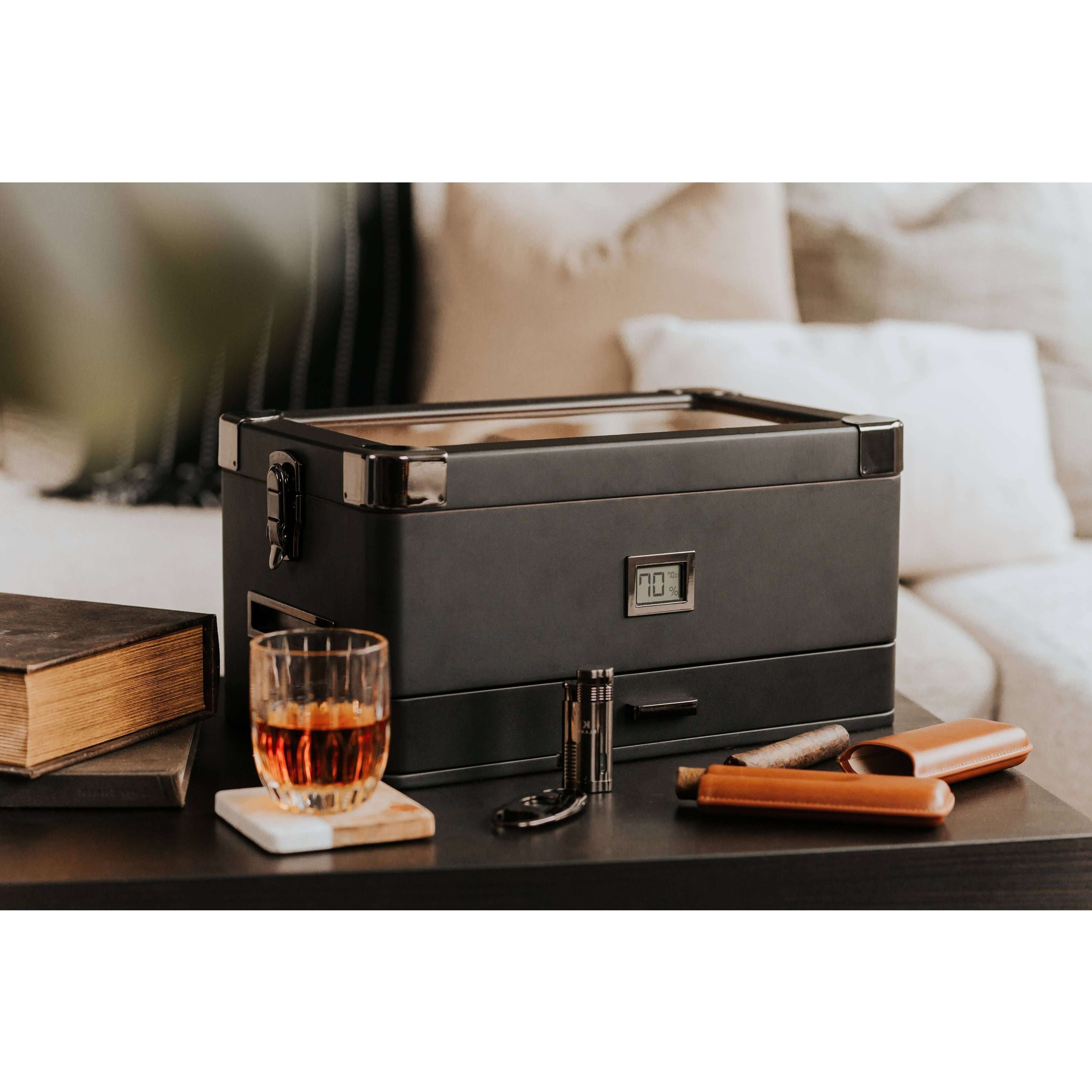 Humidor militaire Black Edition with black matte finish, next to a glass of whiskey and cigar accessories on a table.