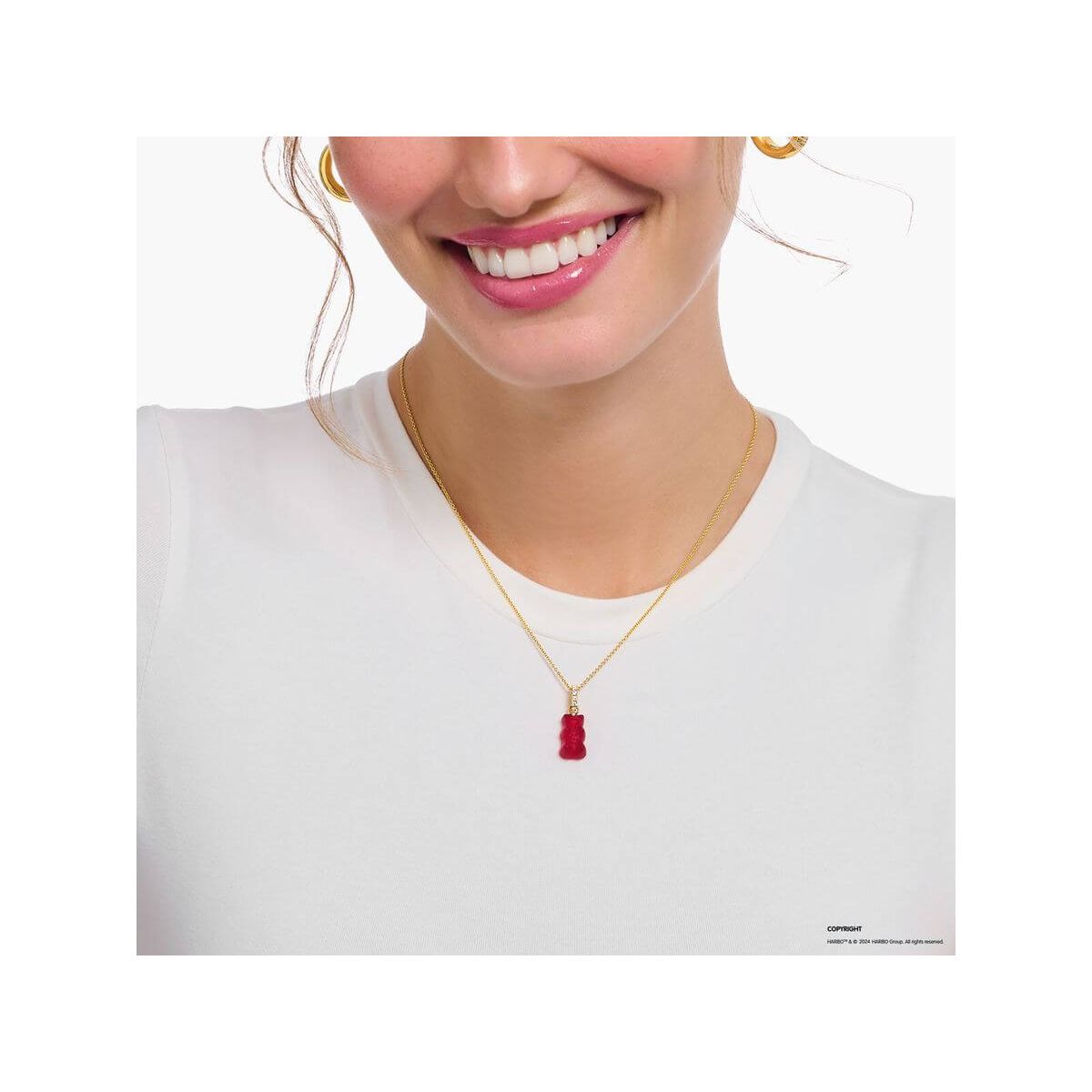 Femme souriante portant une chaîne dorée avec une breloque ours gélifié rouge en cristal, sur un t-shirt blanc.