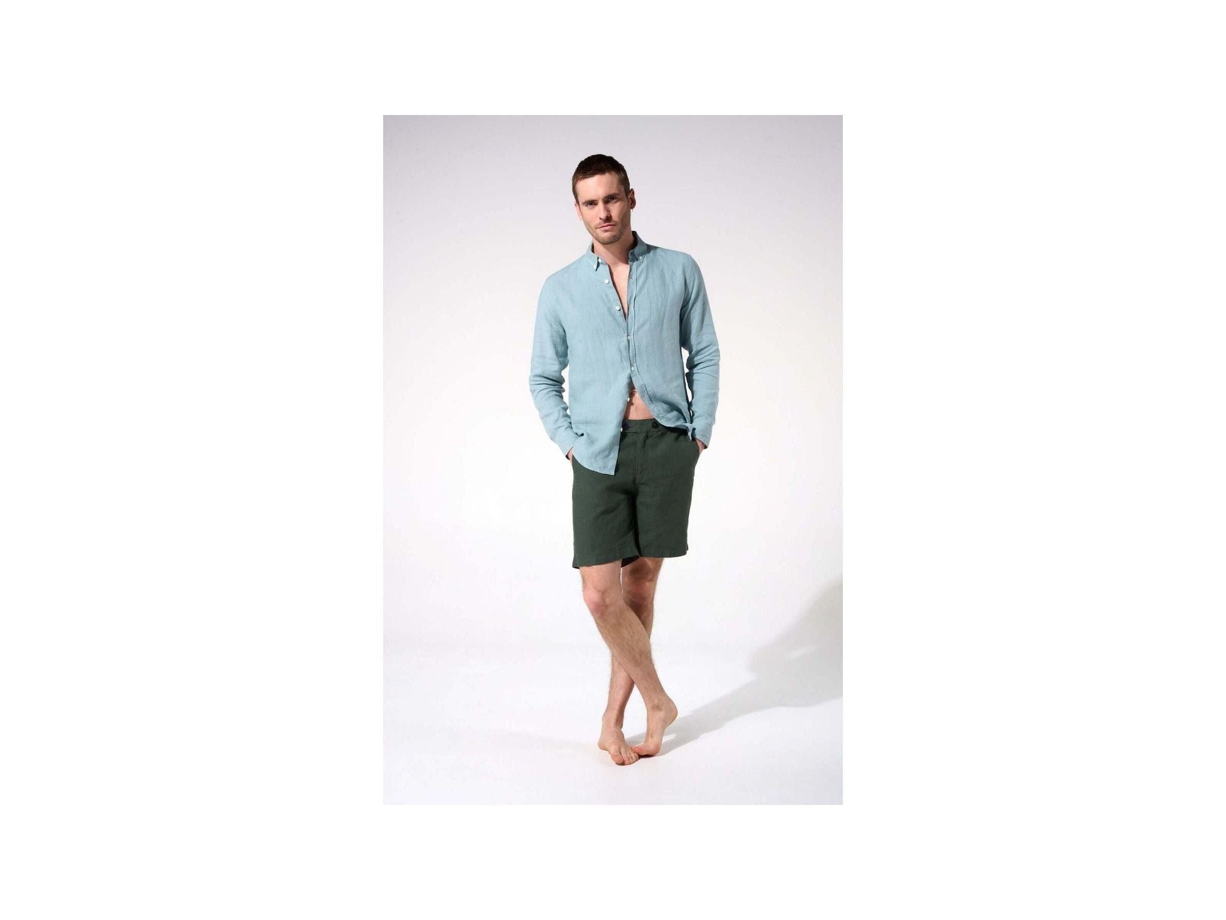 Man wearing a light blue linen shirt and dark green shorts, standing barefoot in a casual pose.