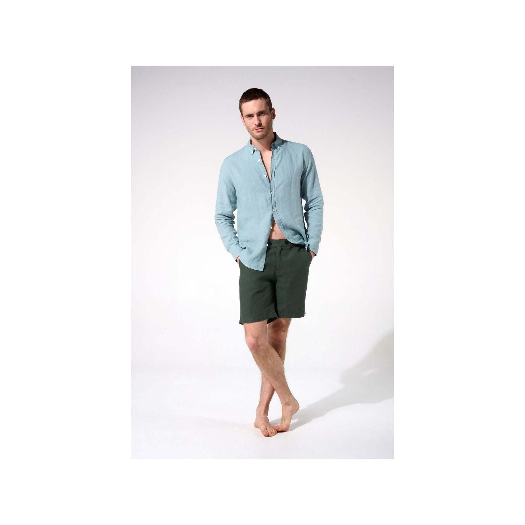 Man wearing a light blue linen shirt and dark green shorts, standing barefoot in a casual pose.