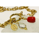 Collier Madone bracelet featuring gold links, red coral bead, and a clear gem on a light stone background.
