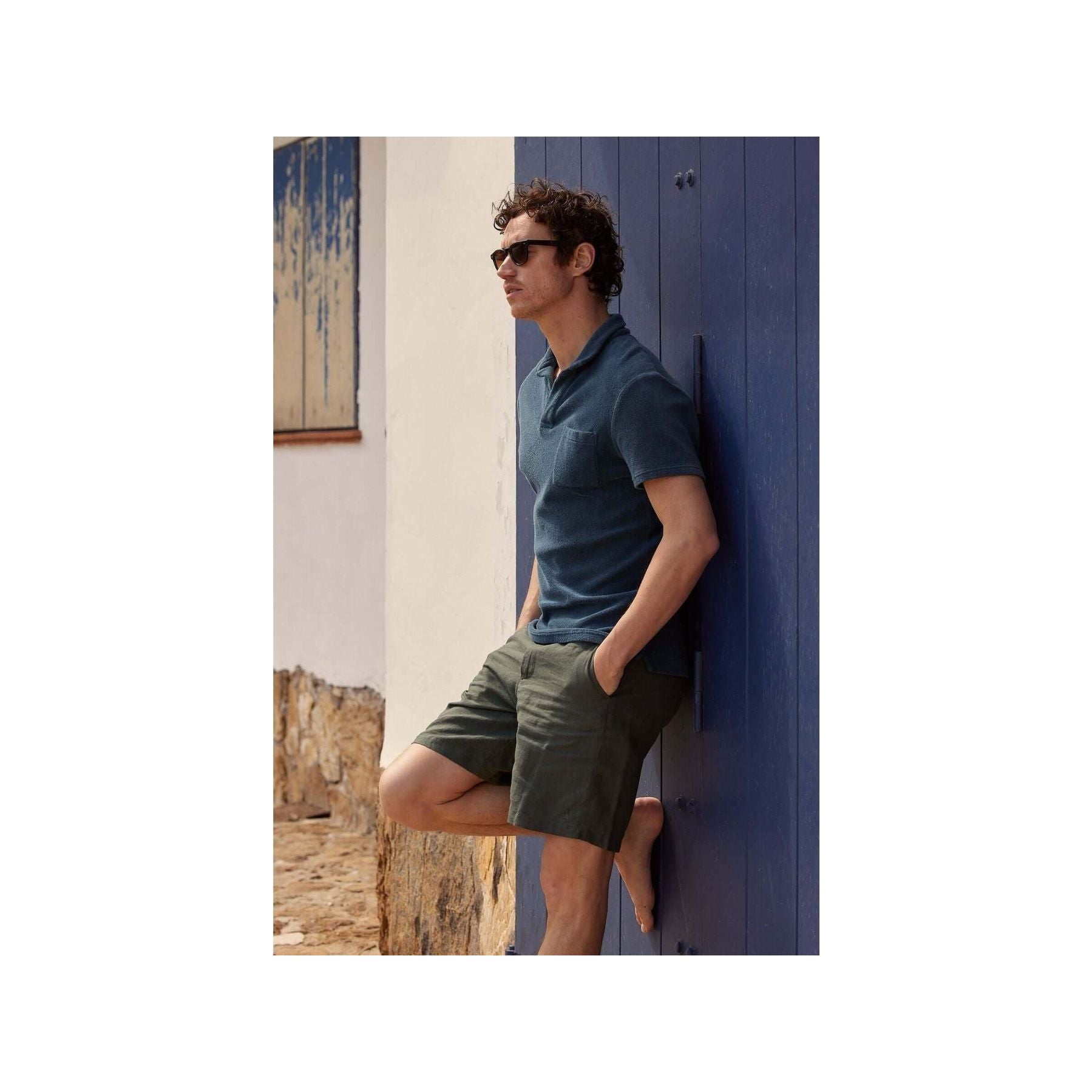 Man wearing Bermuda lin Balsam shorts and a polo shirt, leaning against a blue wall in a stylish summer look.