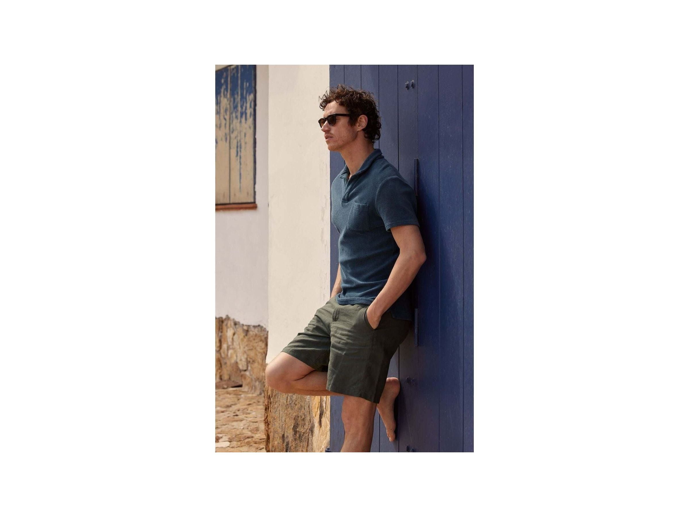 Man wearing Bermuda lin Balsam shorts and a polo shirt, leaning against a blue wall in a stylish summer look.
