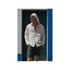 A man wearing a light beige hooded shirt and dark shorts stands in front of a blue and white wall.