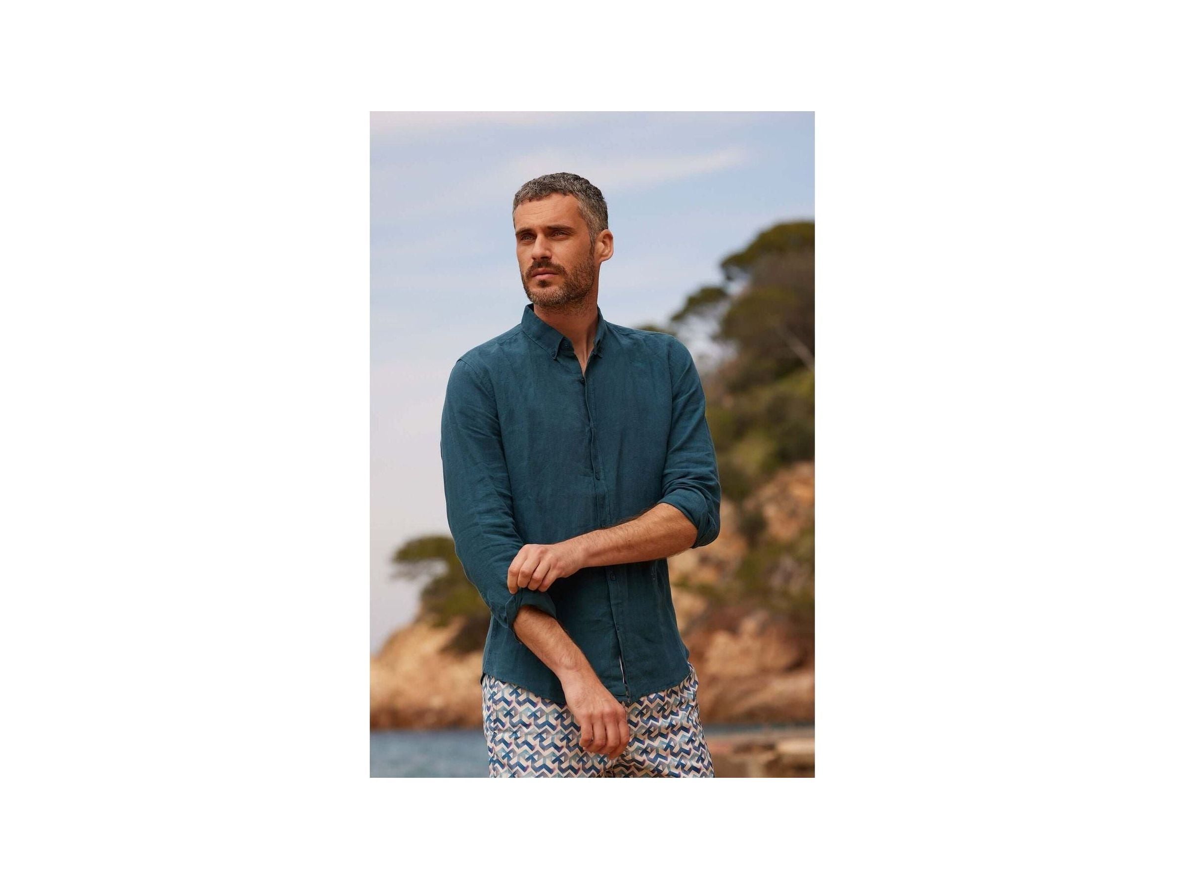 Homme avec chemise en lin bleu, col boutonné, décor naturel, style décontracté en bord de mer.
