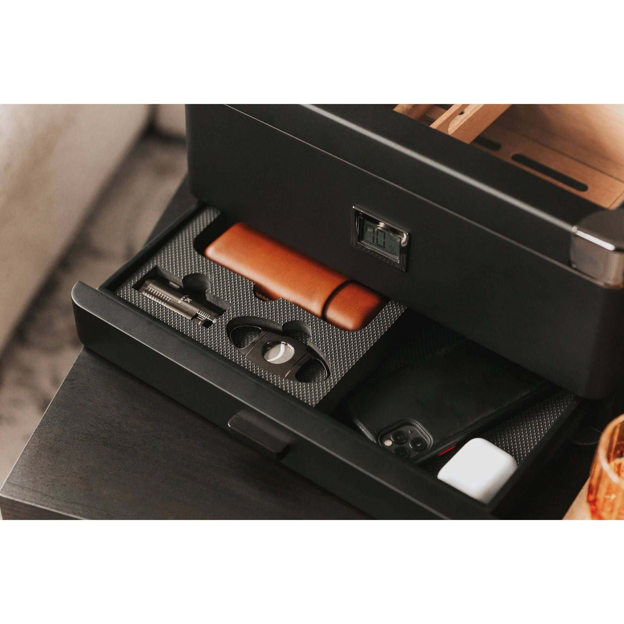 Humidor militaire Black Edition open drawer showcasing accessories like a cigar cutter, leather case, and smartphone.