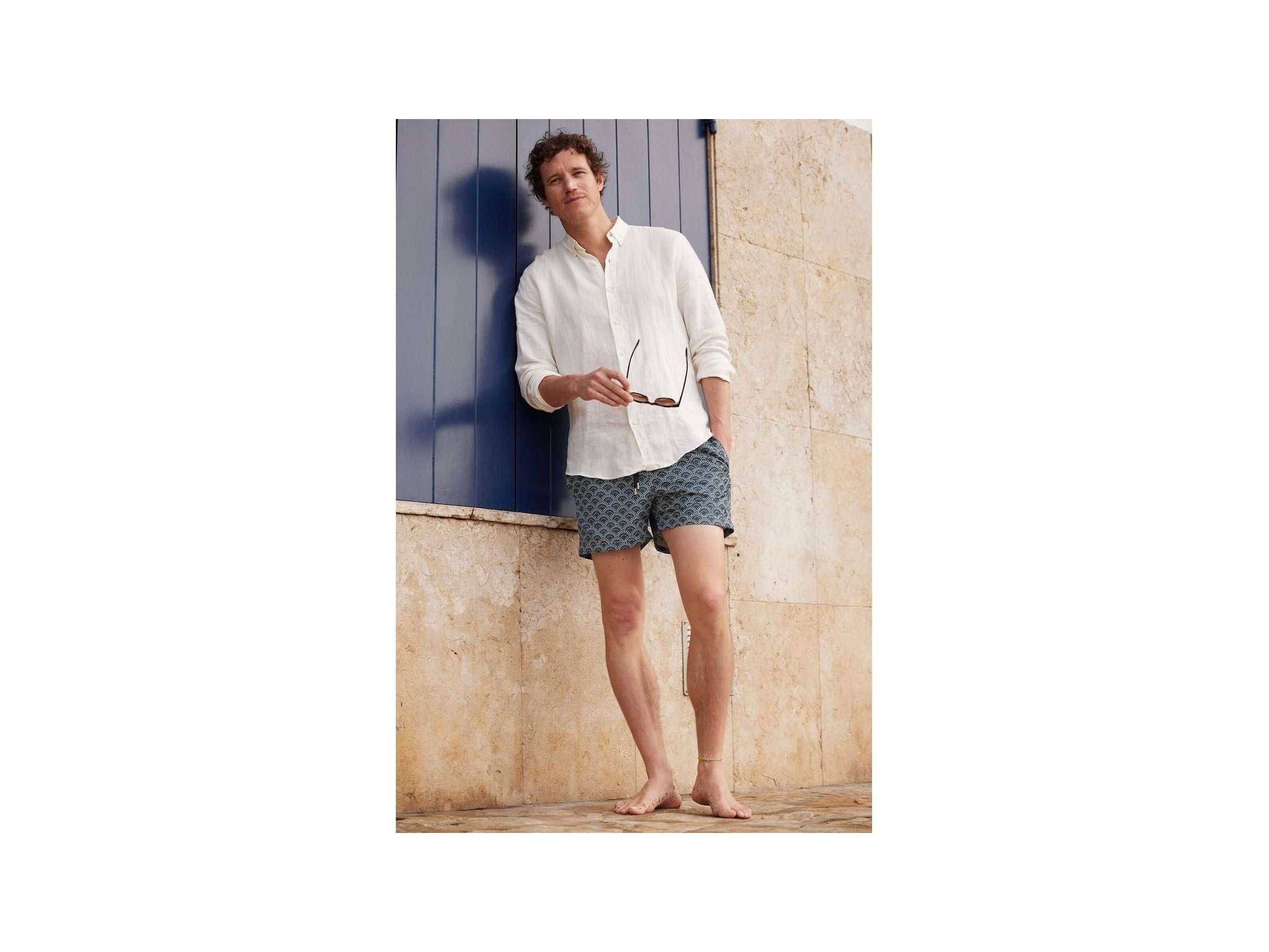 Homme en short de bain Ecume, style décontracté avec chemise blanche, posant devant un mur beige.