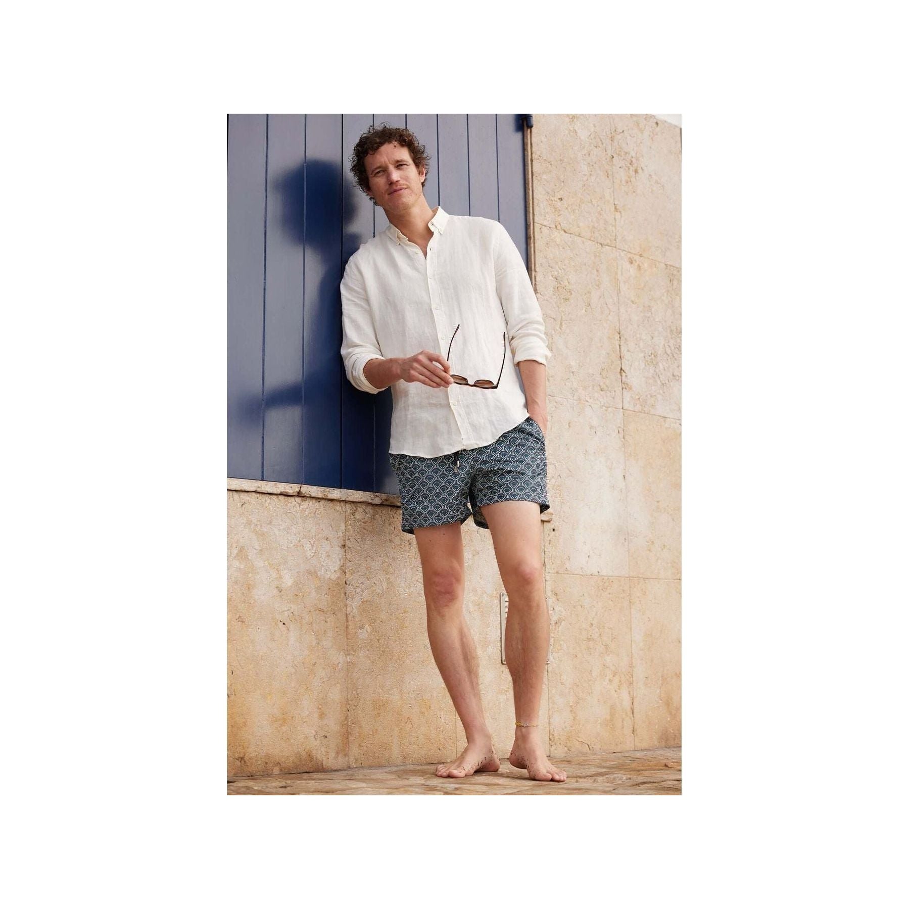 Homme en short de bain Ecume, style décontracté avec chemise blanche, posant devant un mur beige.
