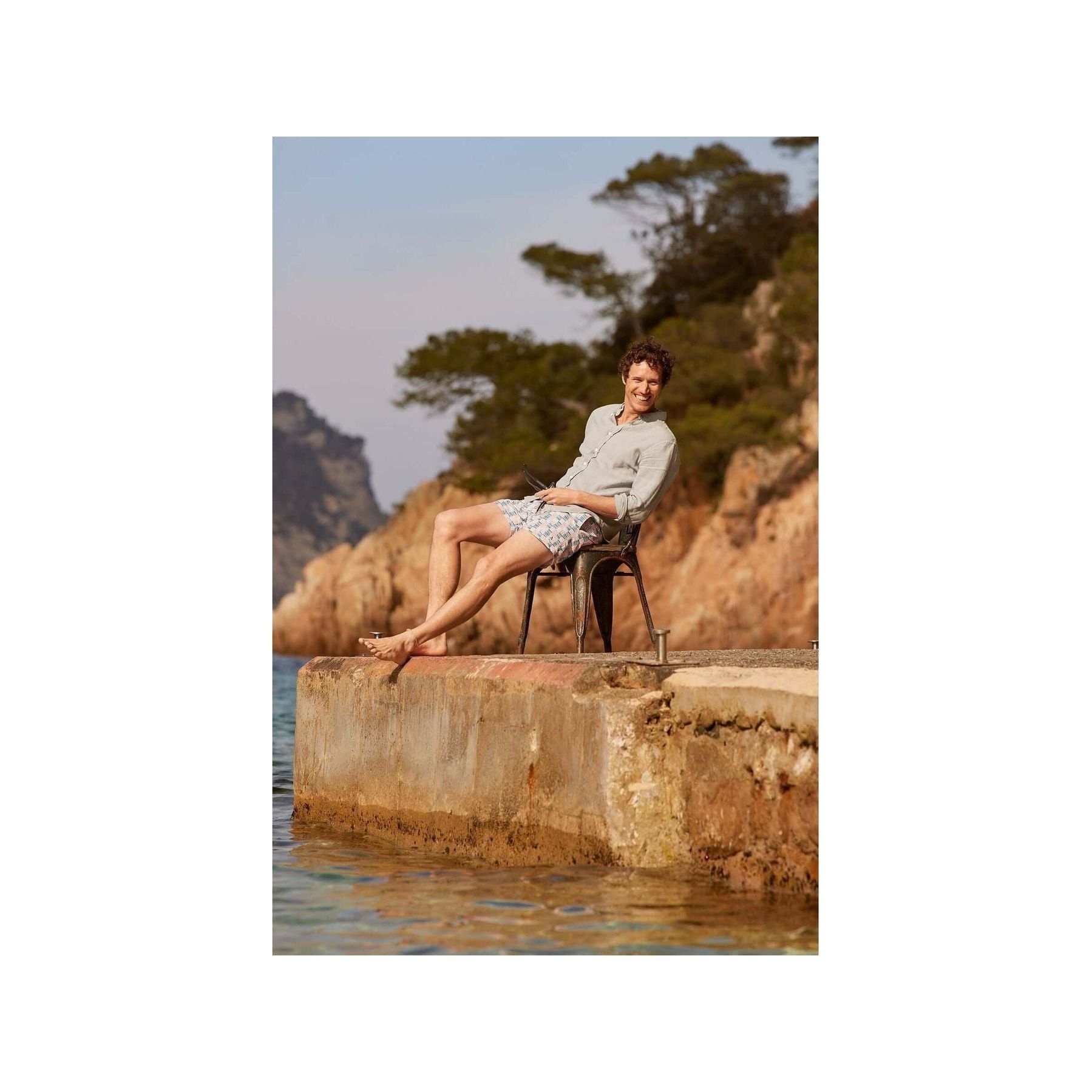 Homme détendu en short de bain Cannage assis sur un quai, en bord de mer, avec des montagnes en arrière-plan.