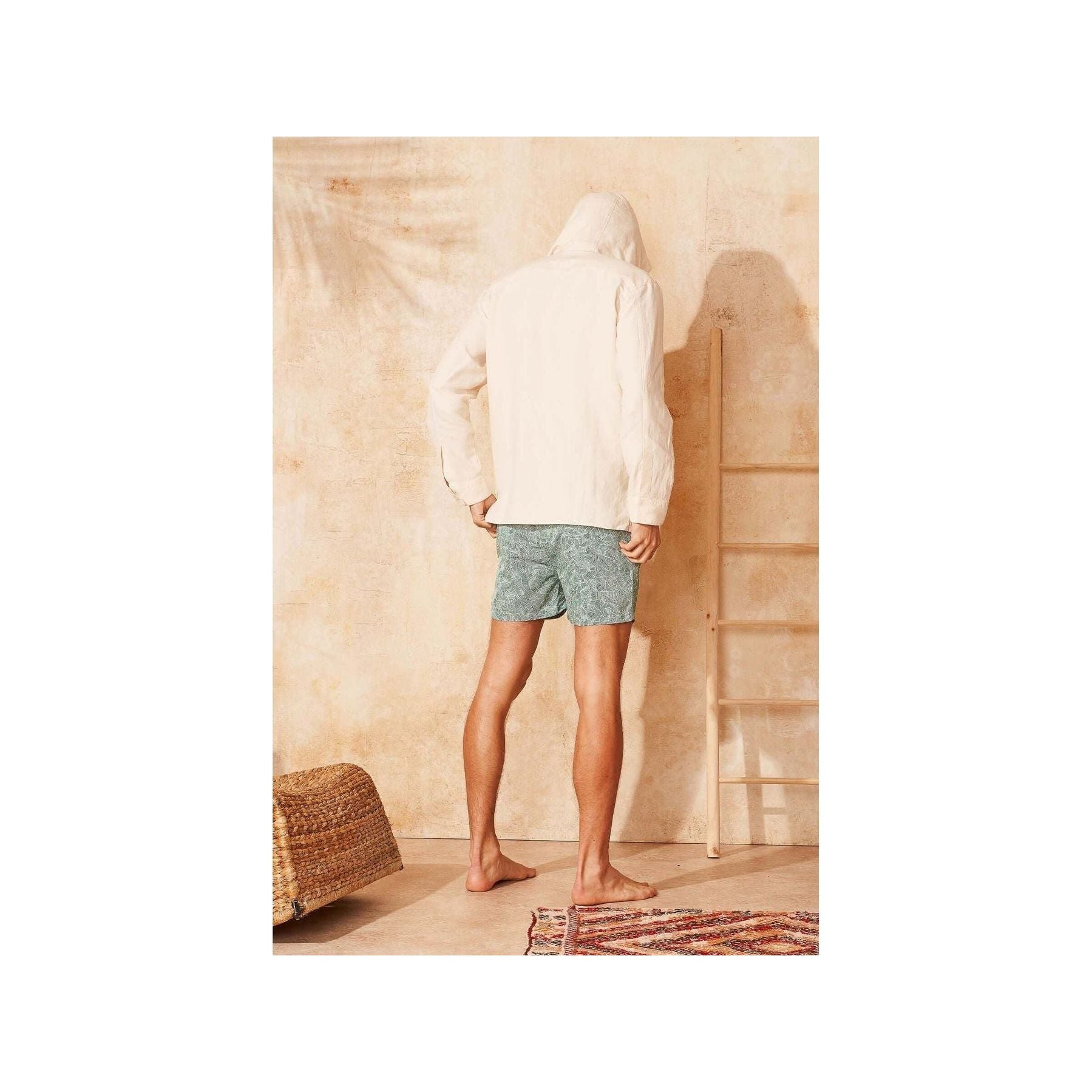 Homme vu de dos portant des shorts de bain Reflets en tissu recyclé, devant un mur beige, avec des pieds nus.
